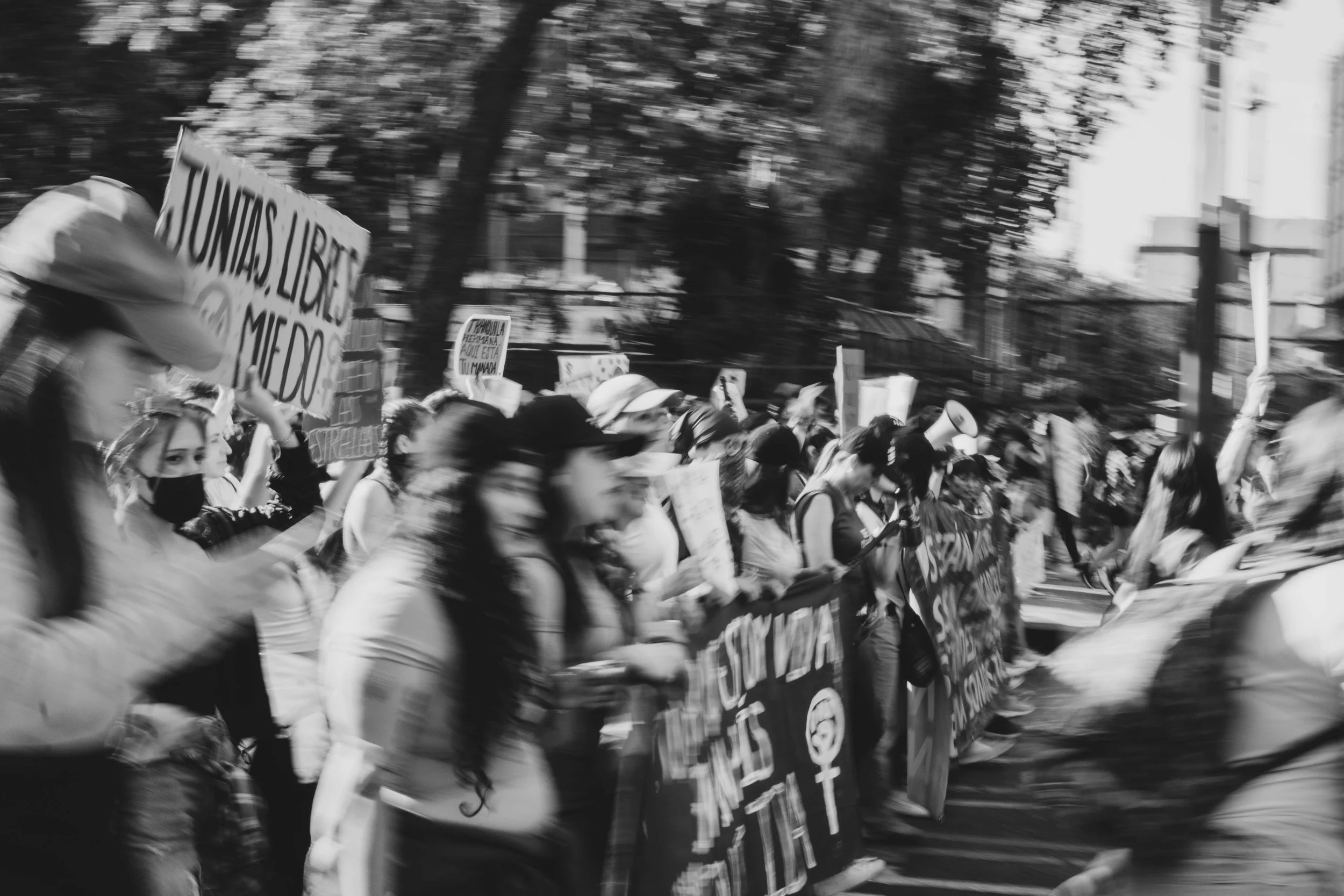 Black and white photograph of a group of people protesting in the street, holding various signs and banners. Some people are wearing face masks and hats.