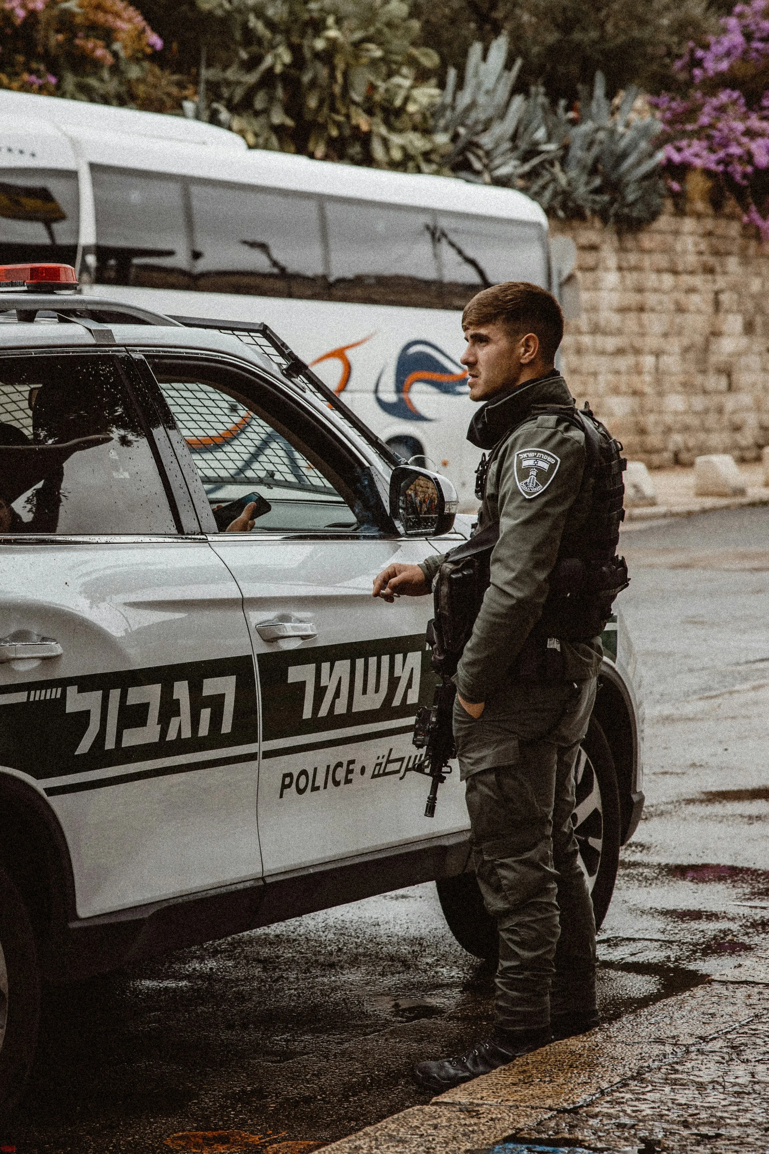 A police officer standing next to a police vehicle on a wet street, with a large bus and greenery in the background.
