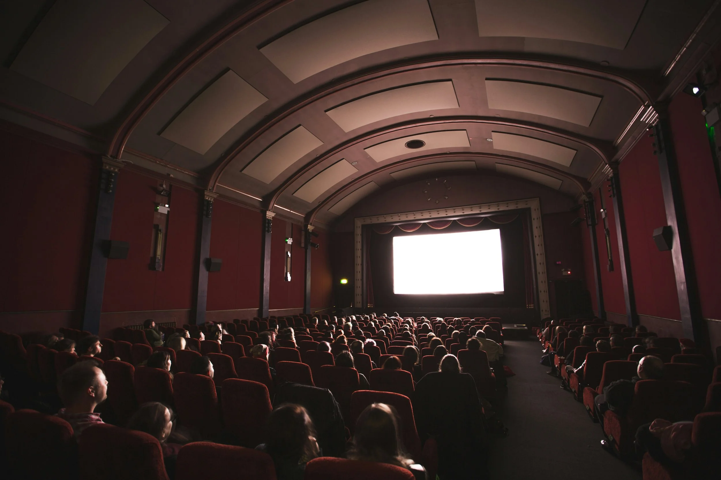 People watching a movie in a theater with a large white screen, red seats, and ornate ceiling.