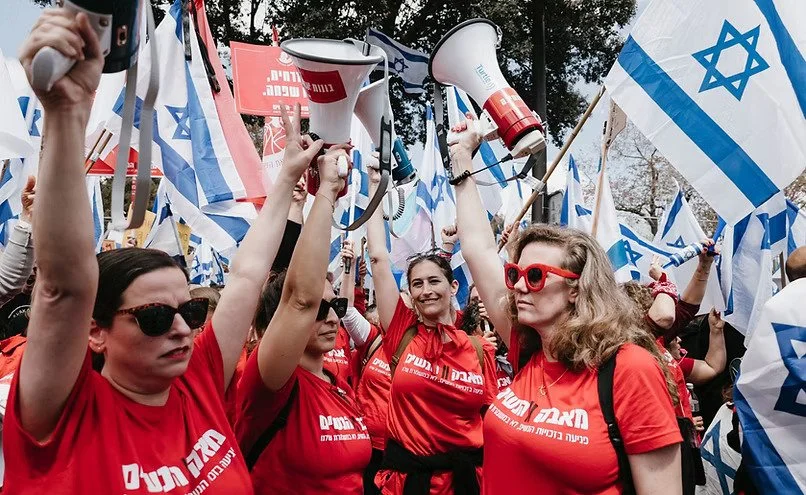A group of women protesting, holding Israeli flags and megaphones, with some wearing red shirts and sunglasses.