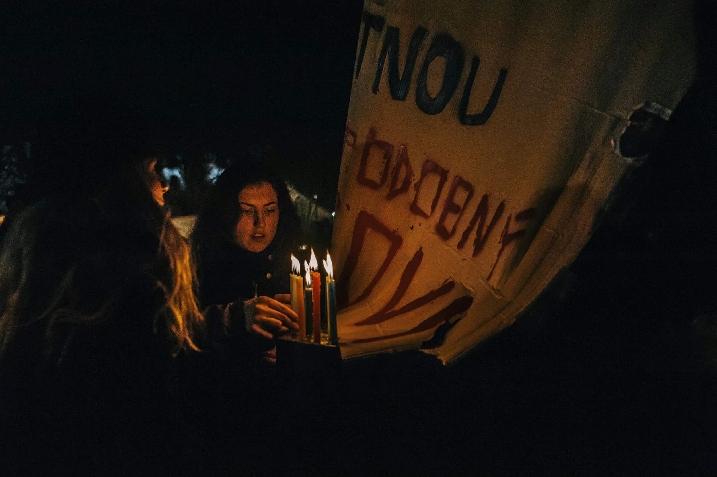 Two women light candles outside at night near a large protest sign with partially visible text.