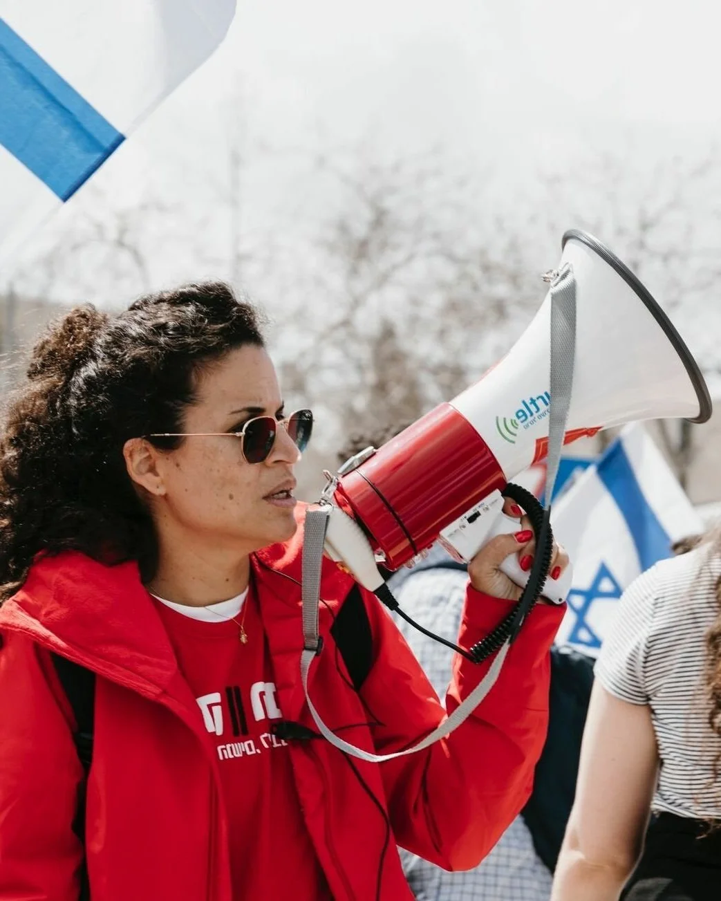 A woman with curly hair, wearing sunglasses and a red jacket, speaking into a megaphone at an outdoor protest or rally.