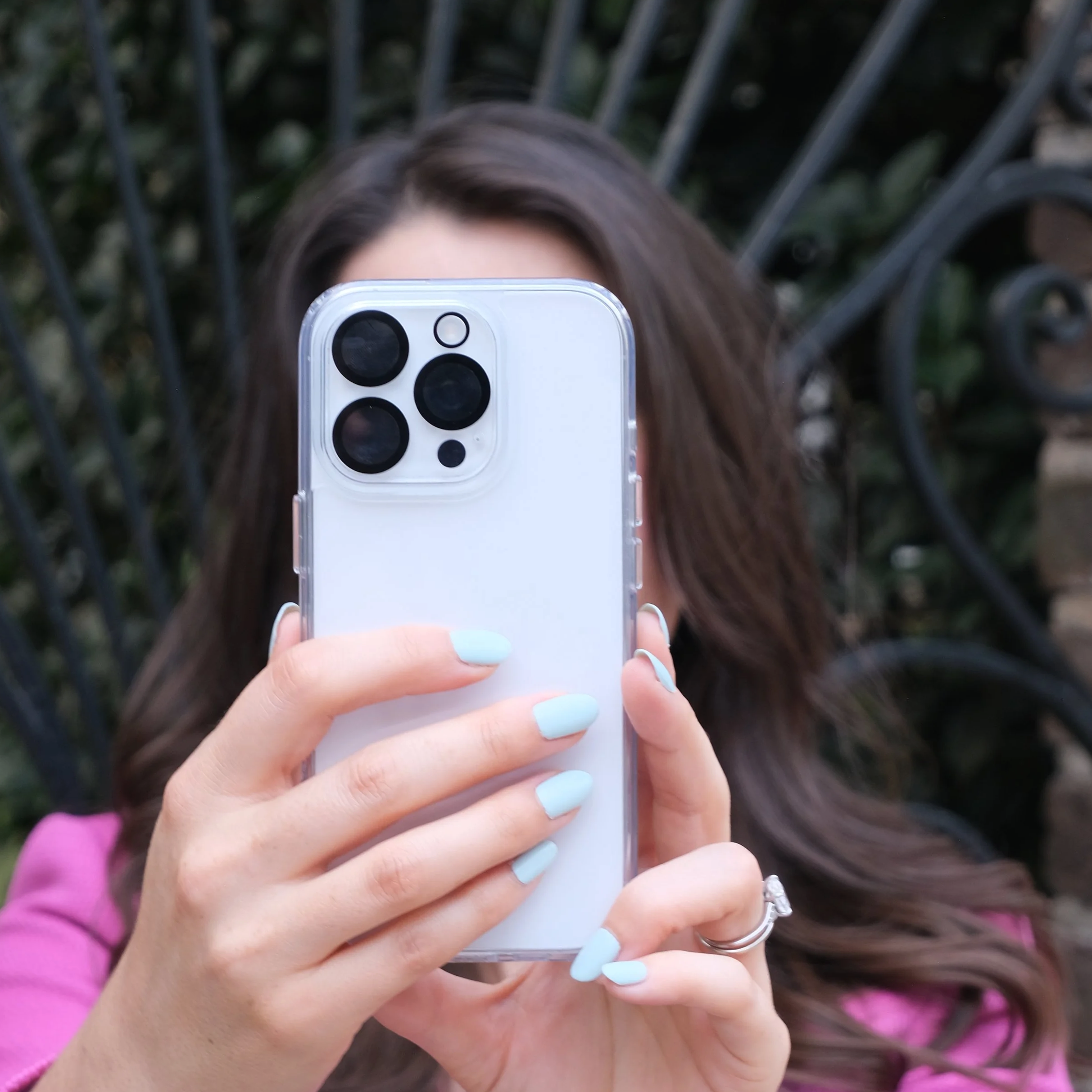 A woman with long brown hair wearing a pink top taking a selfie outdoors with a white iPhone 13 Pro, standing in front of a black wrought iron fence with green foliage behind it.