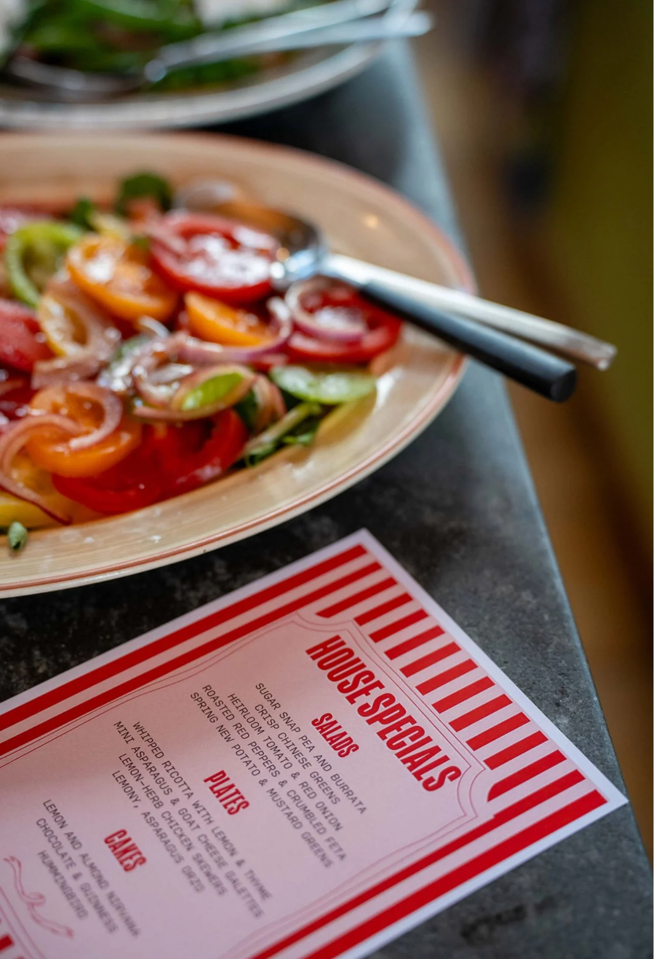A plate of colorful heirloom cherry tomatoes and sliced red onions with a pair of chopsticks resting on the edge of the plate, next to a restaurant menu with a red and white striped border.