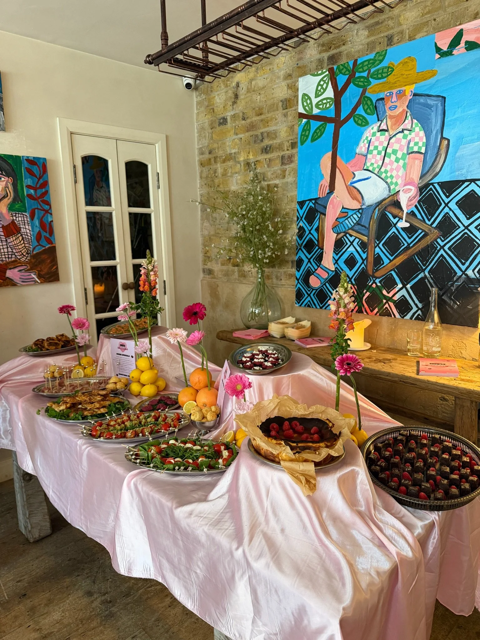 A table set for a celebration with various desserts, snacks, and fruits, decorated with pink flowers in tall vases, and pink tablecloths. In the background, there are colorful paintings on the wall and a rustic, cozy interior.