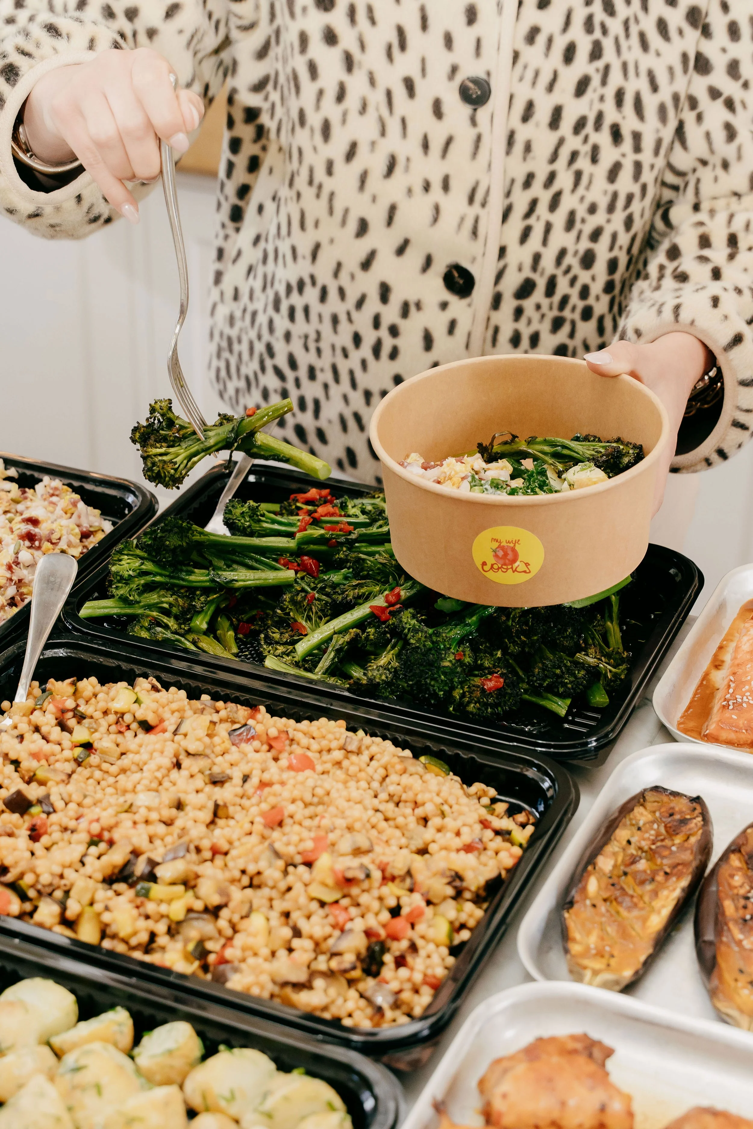 Person wearing a leopard print coat serving themselves food from a buffet with various dishes including roasted vegetables, pasta salad, and egg rolls.