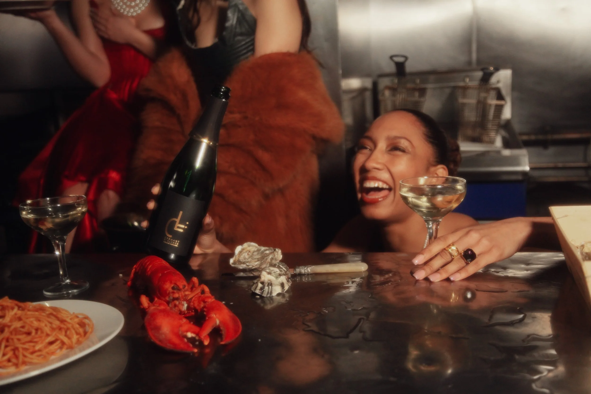 A woman laughing at a bar, with a lobster, spaghetti, and drinks on the counter, holding a bottle of champagne.