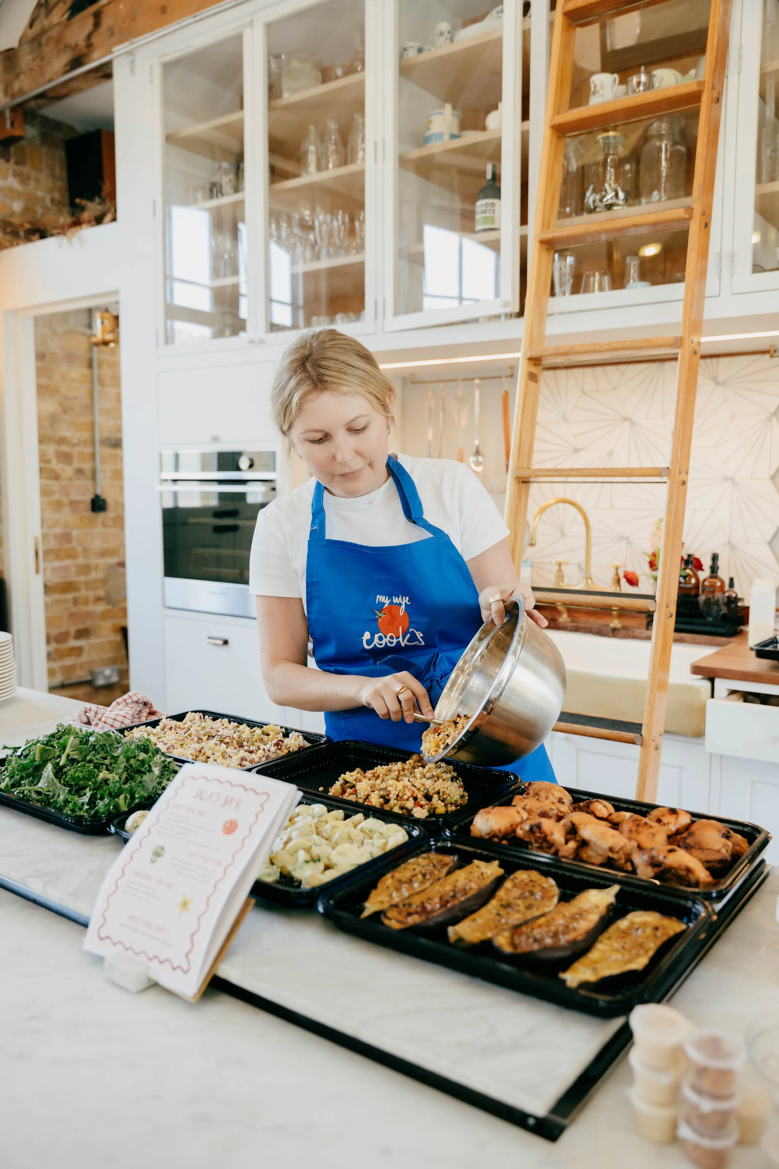 A woman wearing a blue apron serving food at a buffet table with various dishes including greens, grilled chicken, and roasted fish, in a modern kitchen with open cabinets and brick wall.