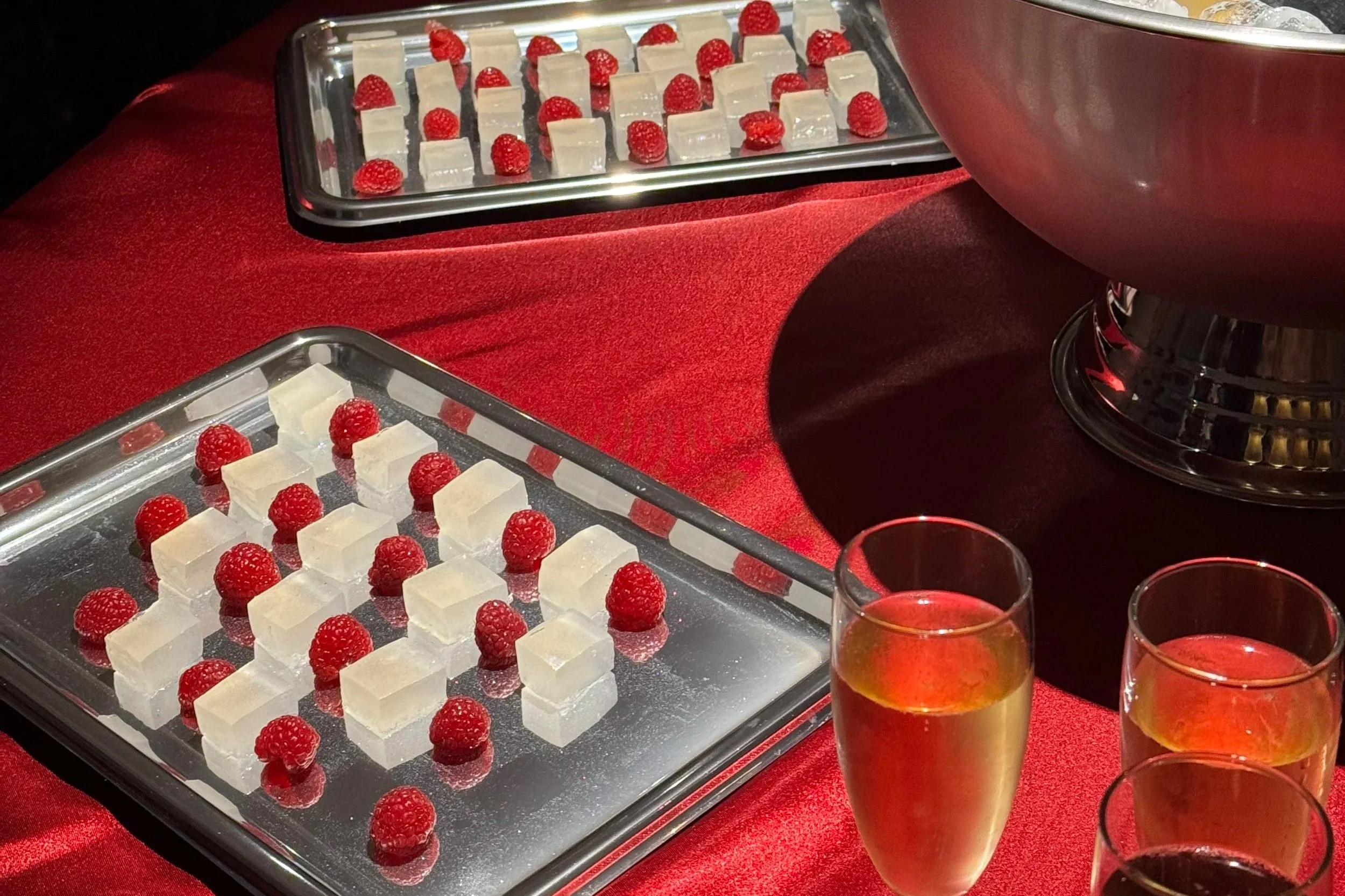 Silver trays with white and red desserts, a large silver bowl, and glasses of champagne and red wine on a red tablecloth.