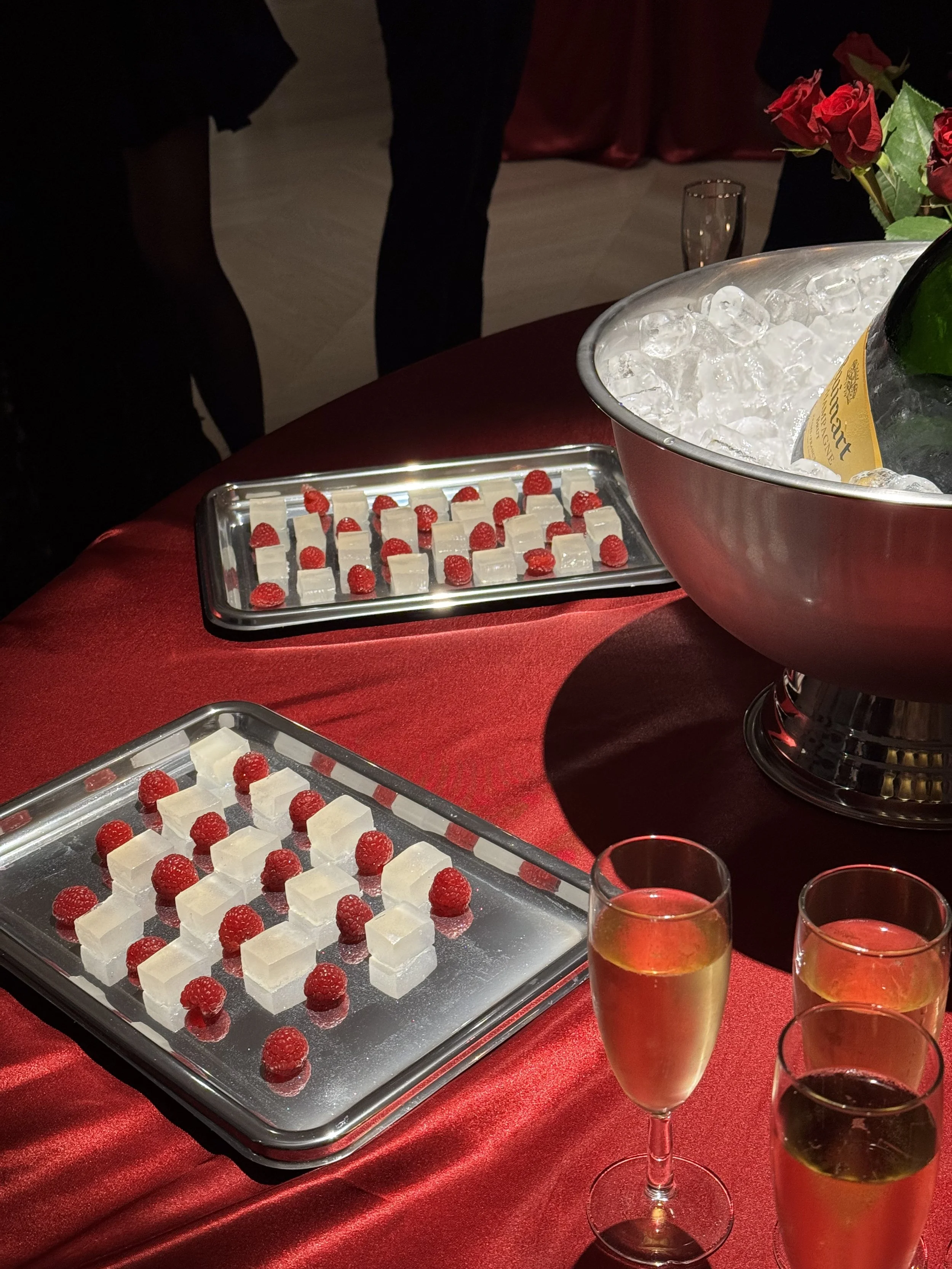  trays of cubed cheese garnished with raspberries, a large bowl of ice with a bottle of champagne, and glasses of champagne on a red tablecloth at a celebration or event.