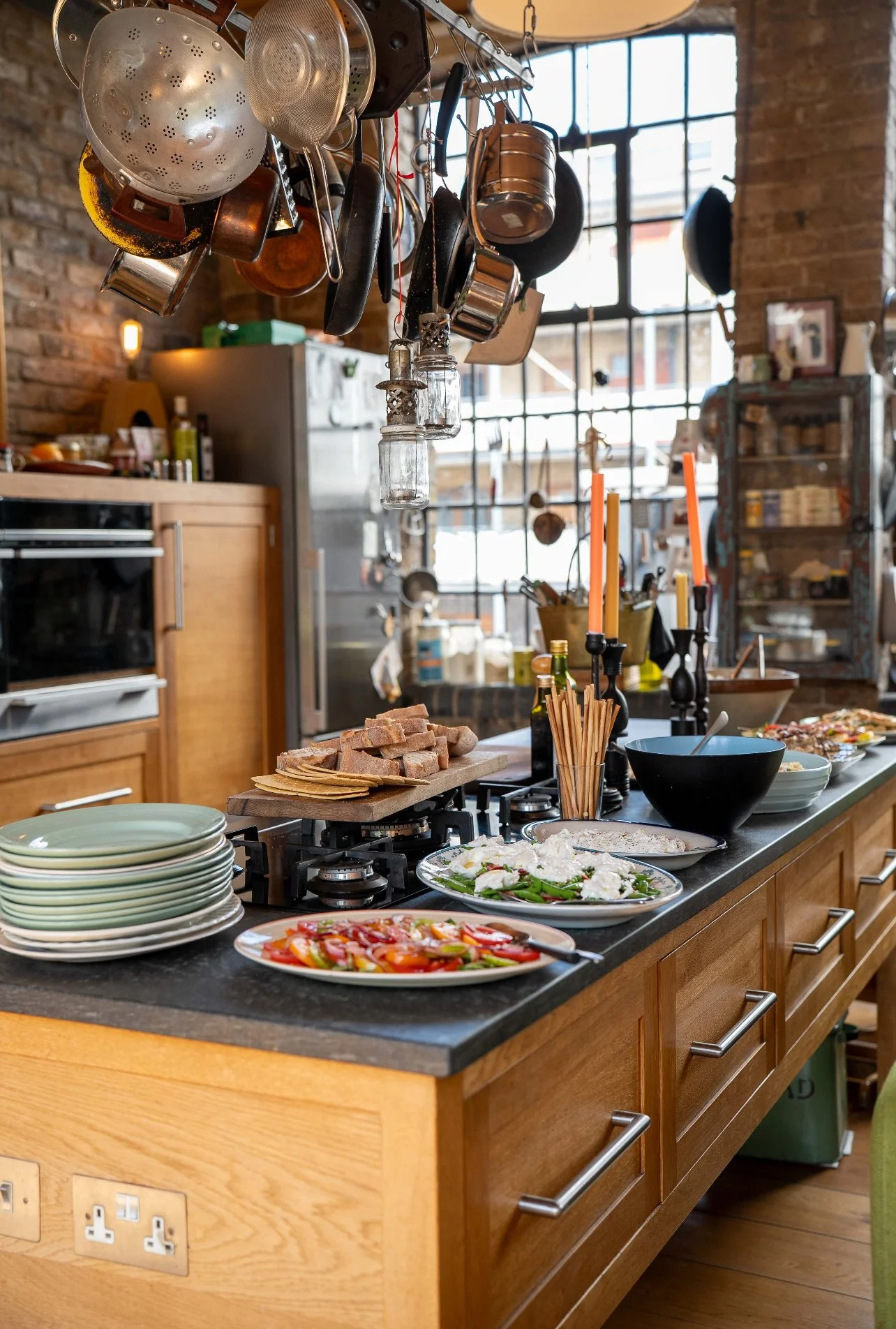 A kitchen with hanging pots and pans above a black countertop island. The island has plates of food, bread, and utensils with a rustic brick wall and large window in the background.