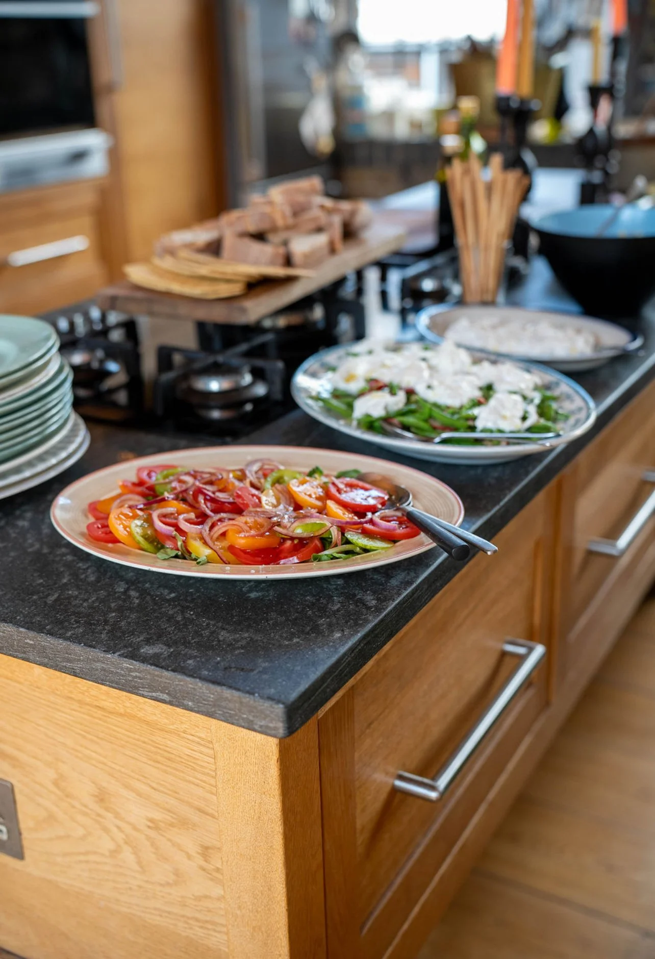 A buffet-style spread of fresh vegetables and salads on a kitchen counter, including sliced tomatoes and peppers, with plates of cooked meats and other dishes in the background.