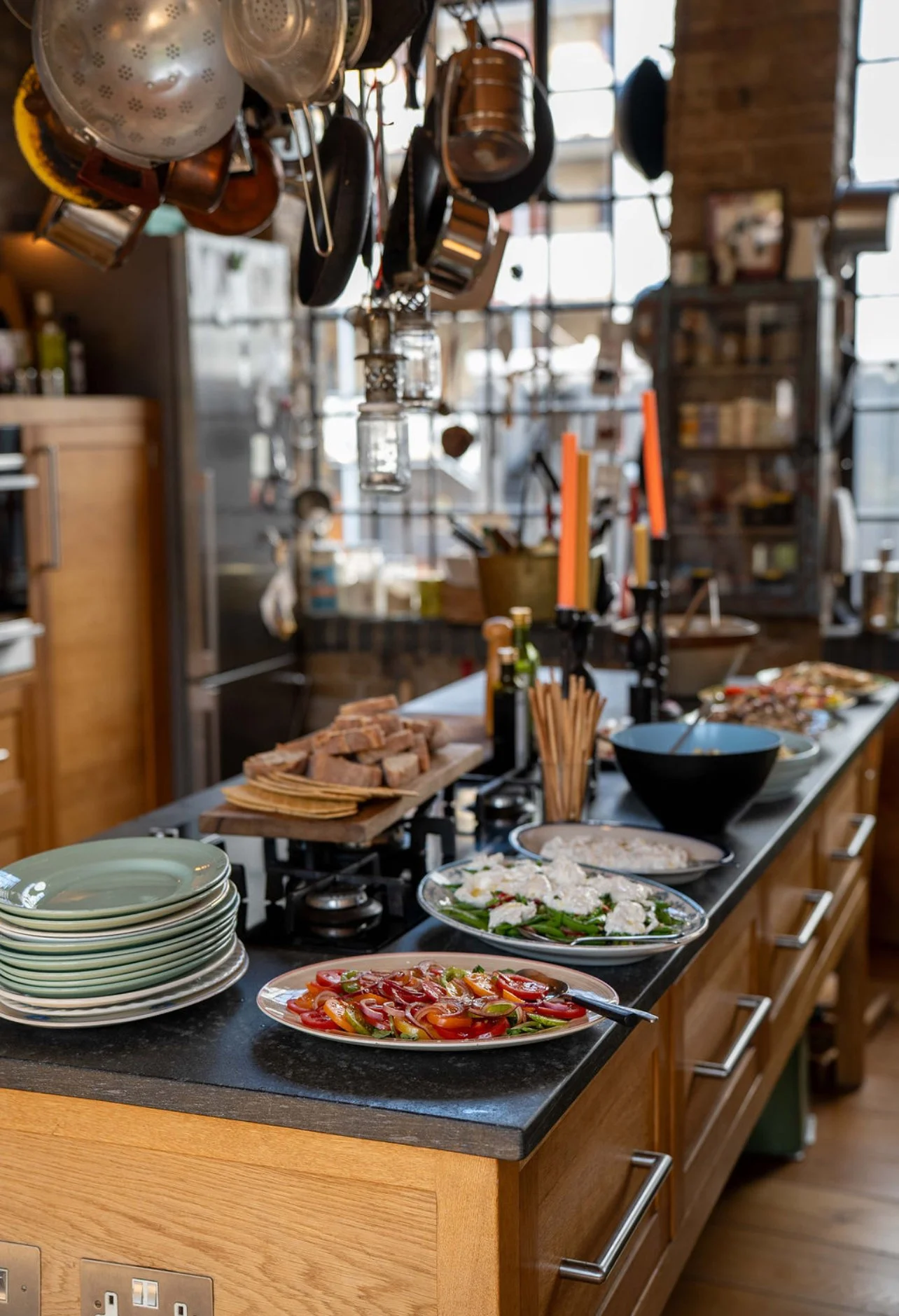 A kitchen countertop with sliced tomatoes and other vegetables, stacked plates, bread, and various dishes, with hanging pots and kitchen supplies in the background.
