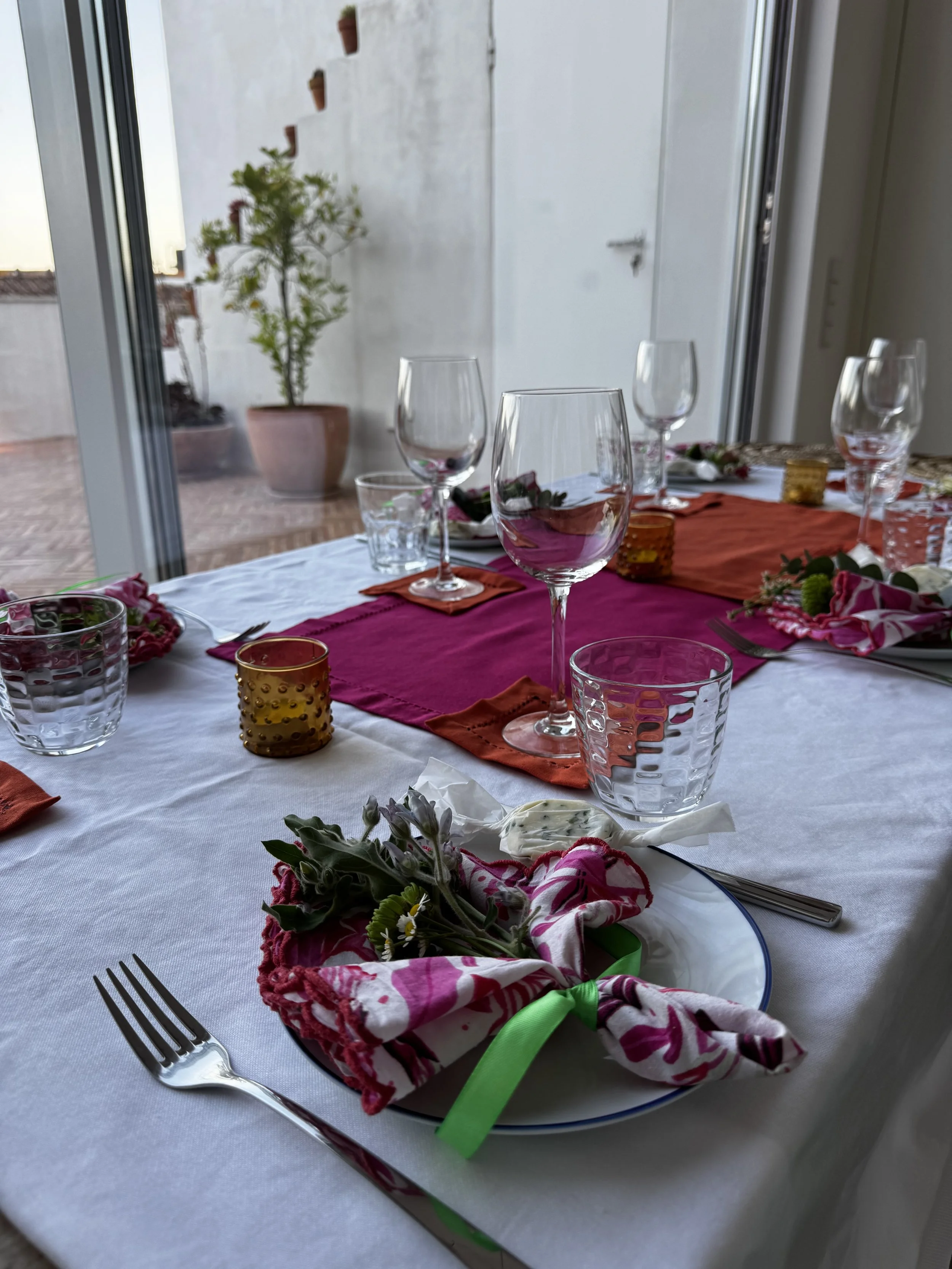 A decorated dining table set for a meal with wine glasses, water glasses, a floral napkin with a green ribbon on a plate, and colorful placemats, in a room with large windows and potted plants outside.