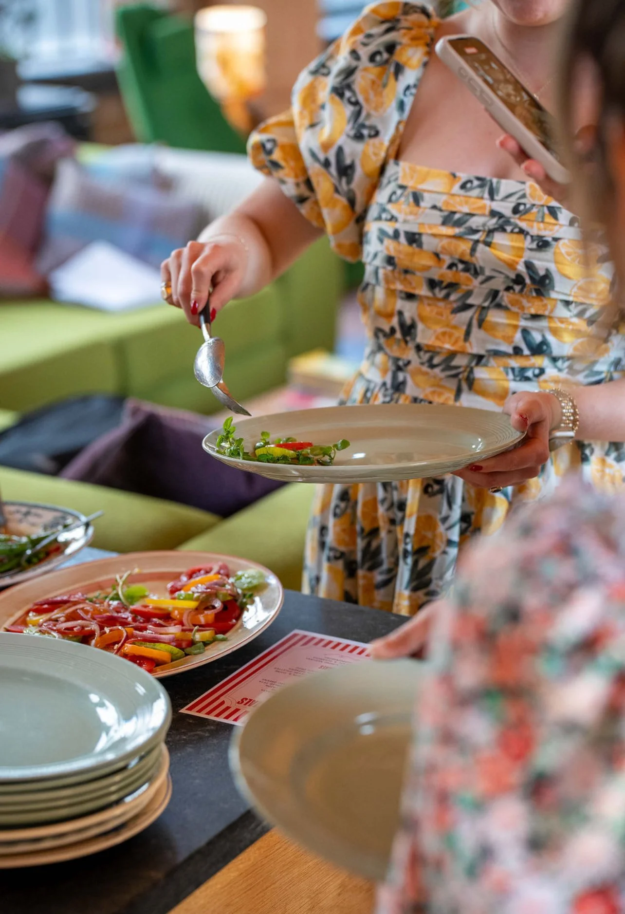 Person serving salad onto a plate at a buffet; plates of salad and other dishes on the table.