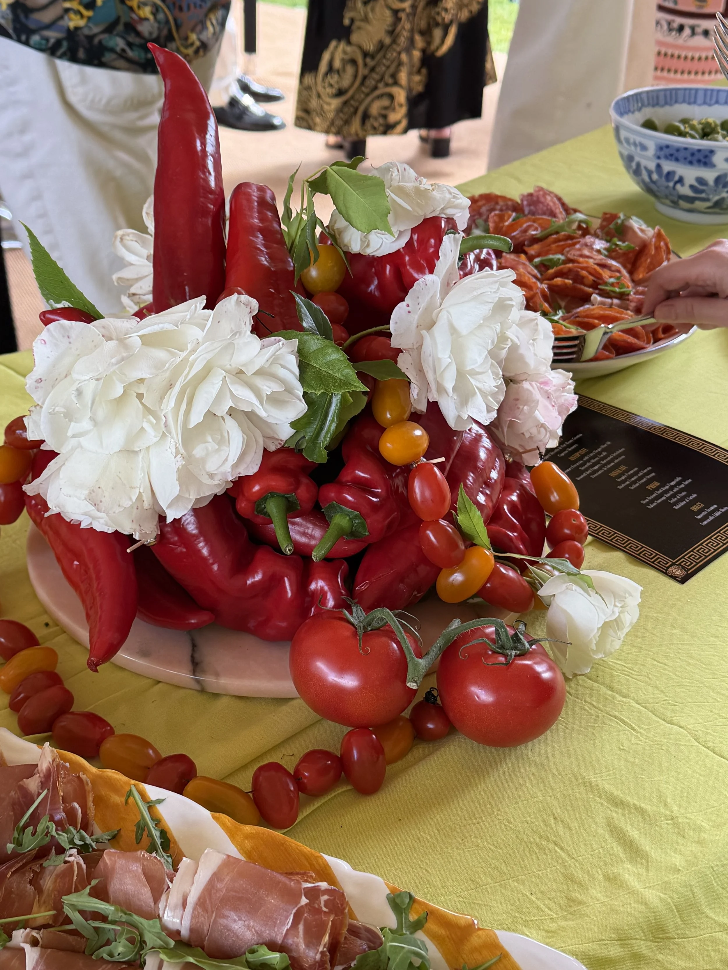 Close-up of a sushi platter with tomatoes, red chili peppers, and white flowers on a yellow tablecloth at a buffet or party table.