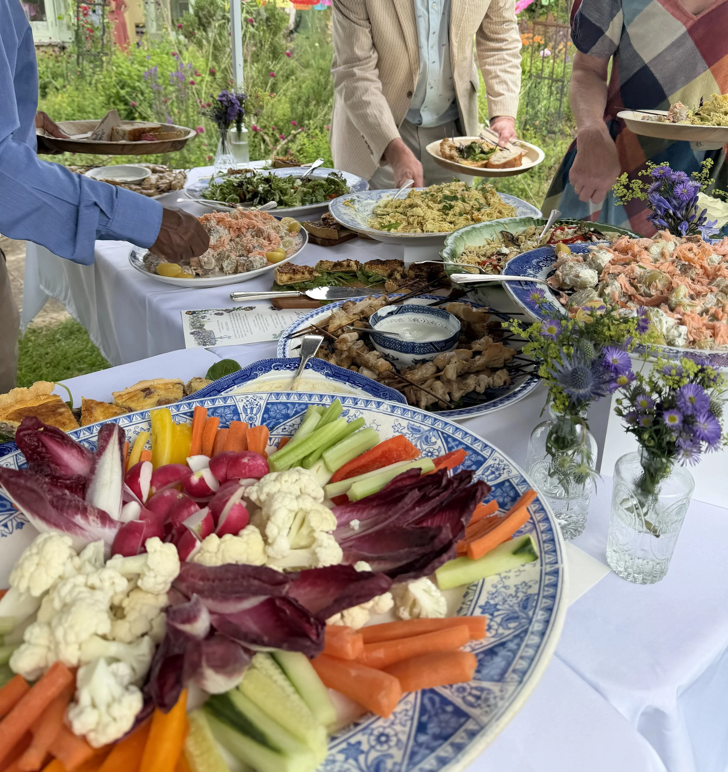 A table set outdoors with various dishes including salads, vegetables, and cooked foods, surrounded by colorful flowers and guests serving themselves from the buffet.