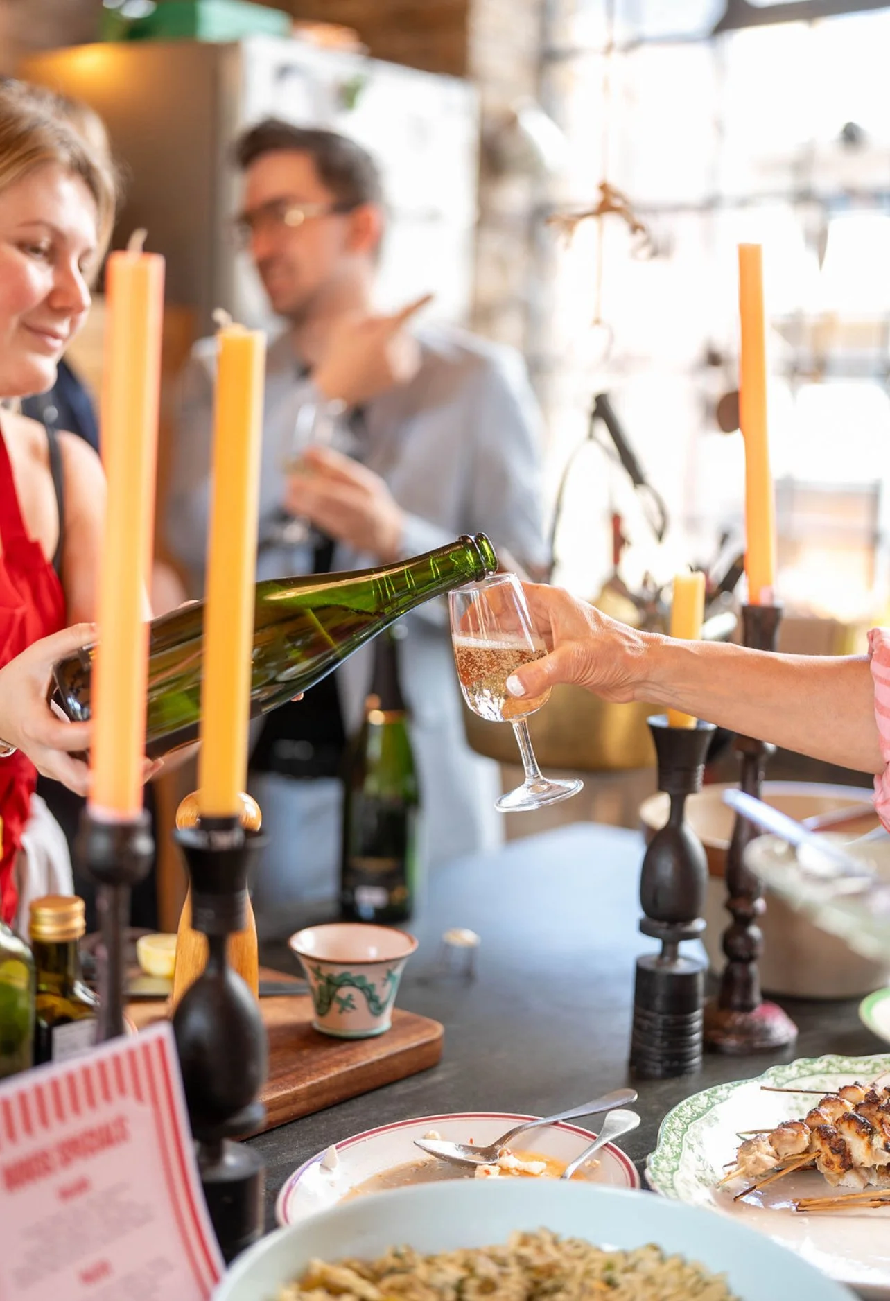 Someone pouring champagne into a glass during a gathering with food and candles on the table.