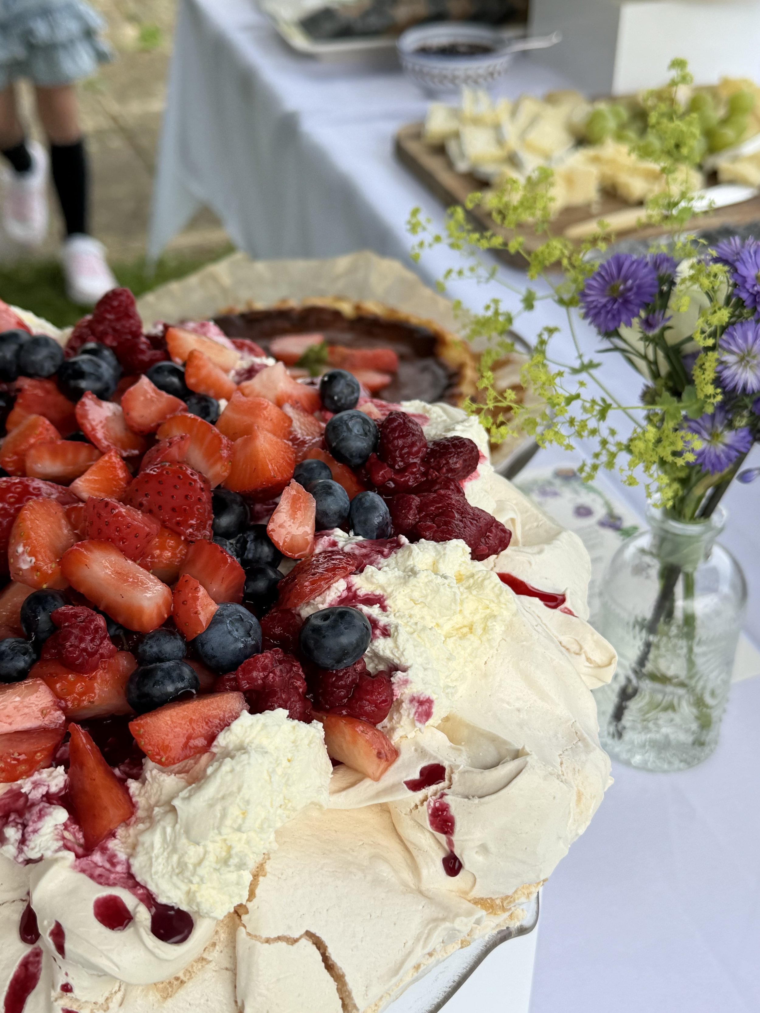 Close-up of a berry-topped pavlova with whipped cream, on a white table with flowers in a glass vase in the background.