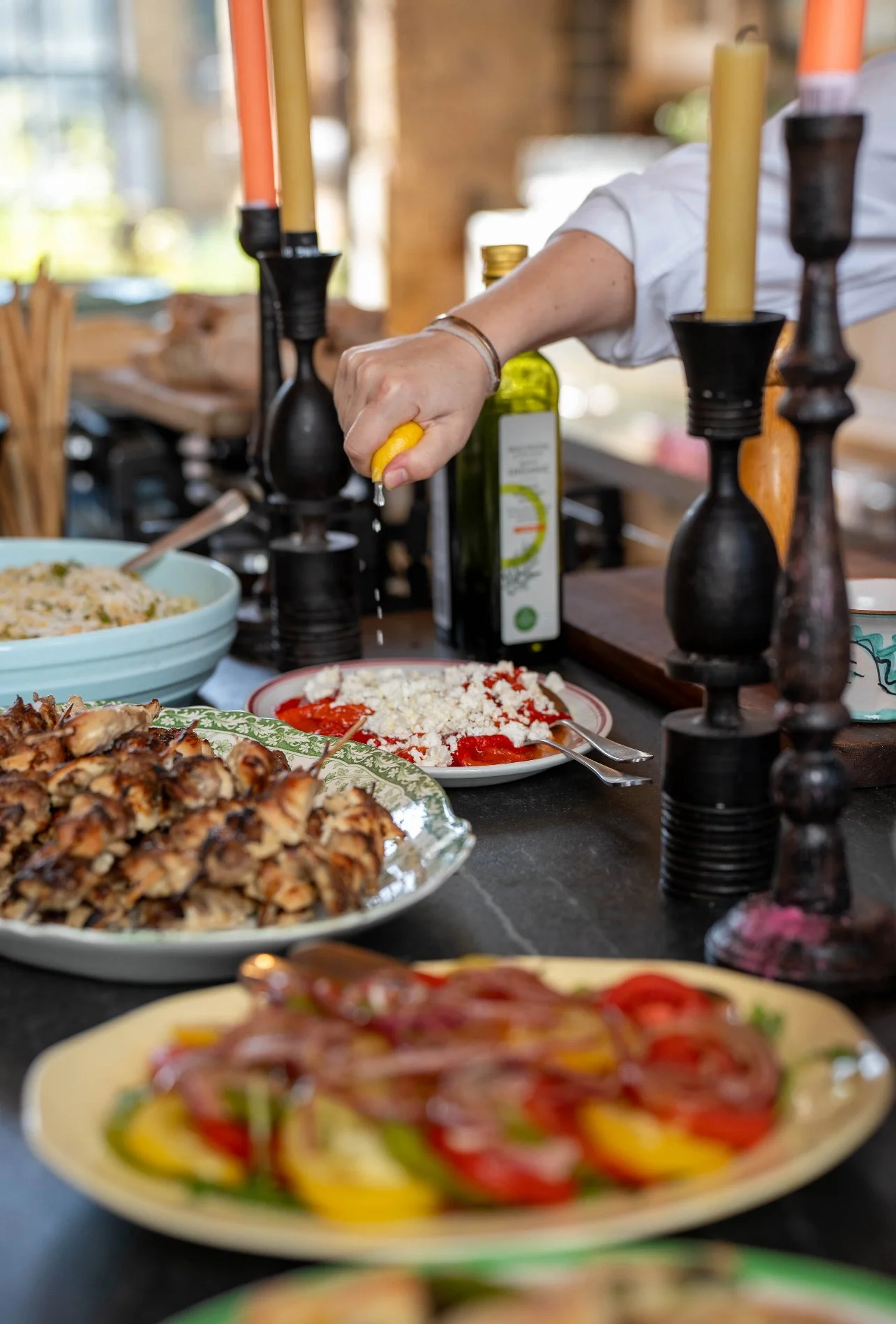 Person squeezing lemon over a platter of crumbled feta cheese and sliced tomatoes on a plate, with other dishes and candles on the counter.