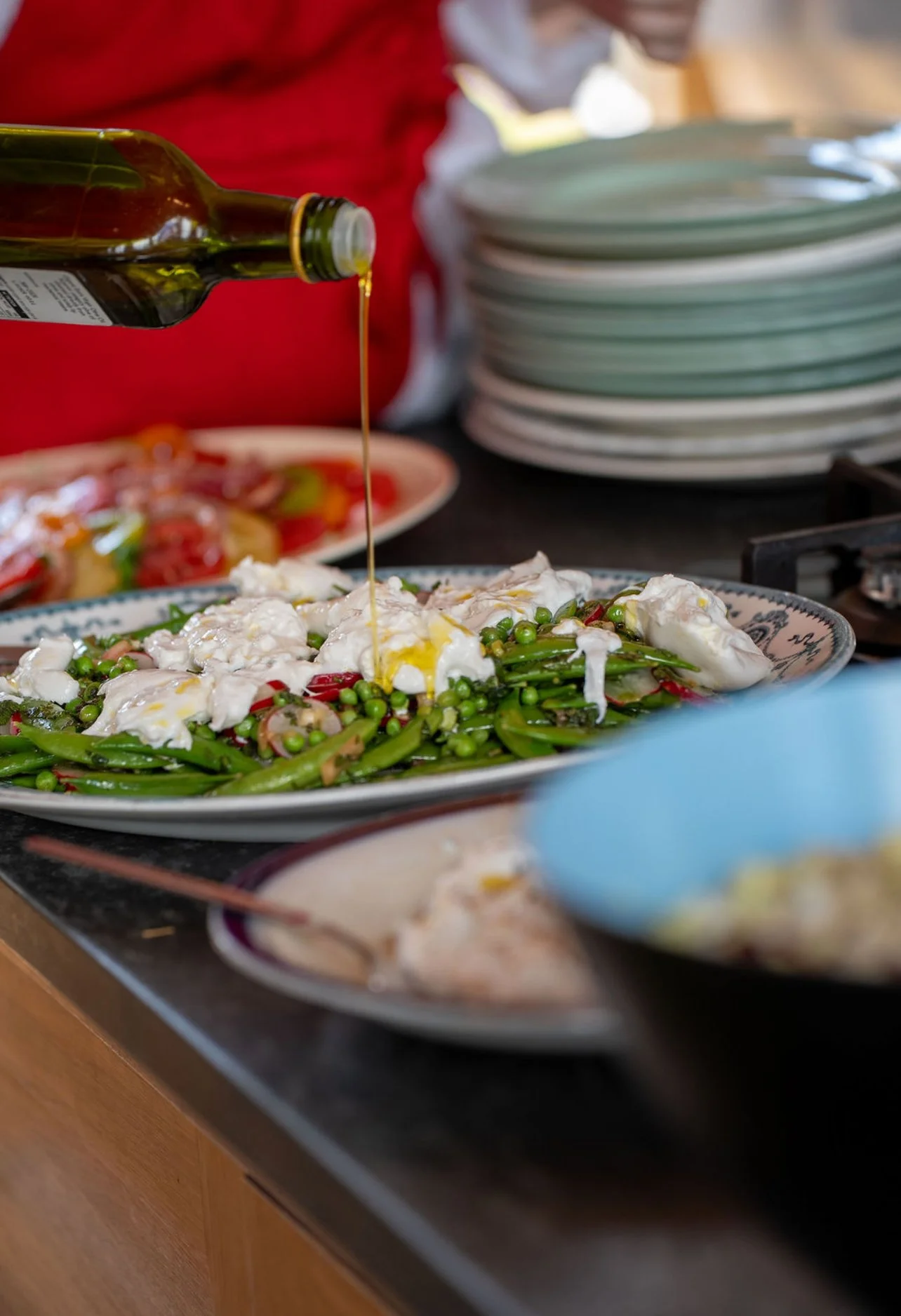 A plate of salad with green beans, snap peas, and dollops of white cheese, topped with olive oil being poured from a green bottle. In the background, there are stacked plates and people serving food.