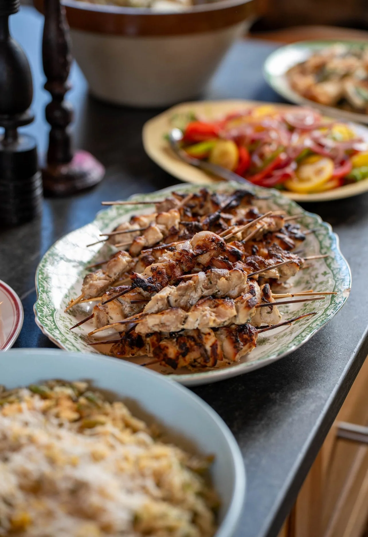 Plate of grilled chicken skewers on a decorative plate, with other dishes of pasta and salad in the background, on a dark table.