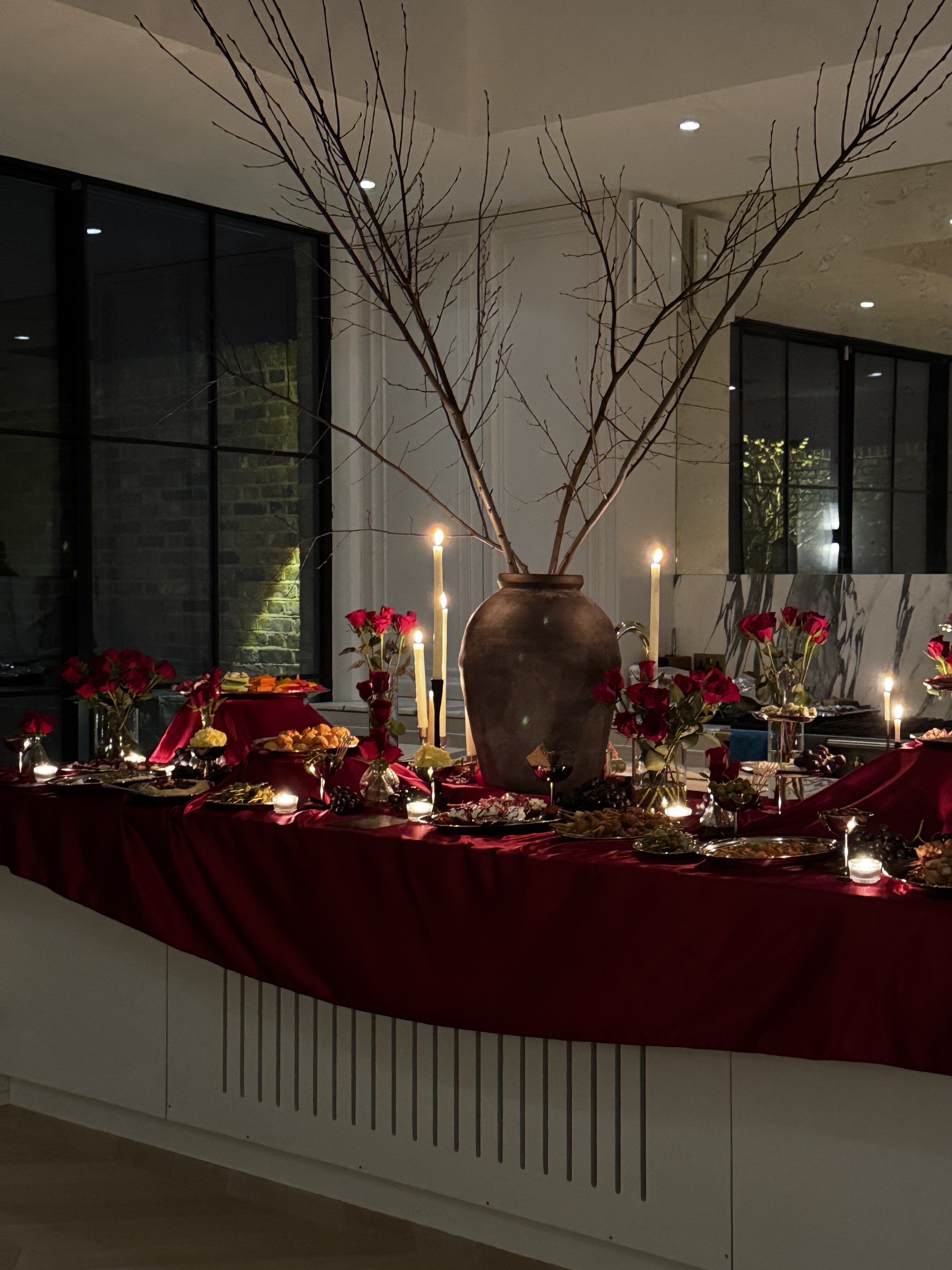 A dinner table decorated with red flowers, candles, and a large brown vase with bare branches, set near large windows at night.