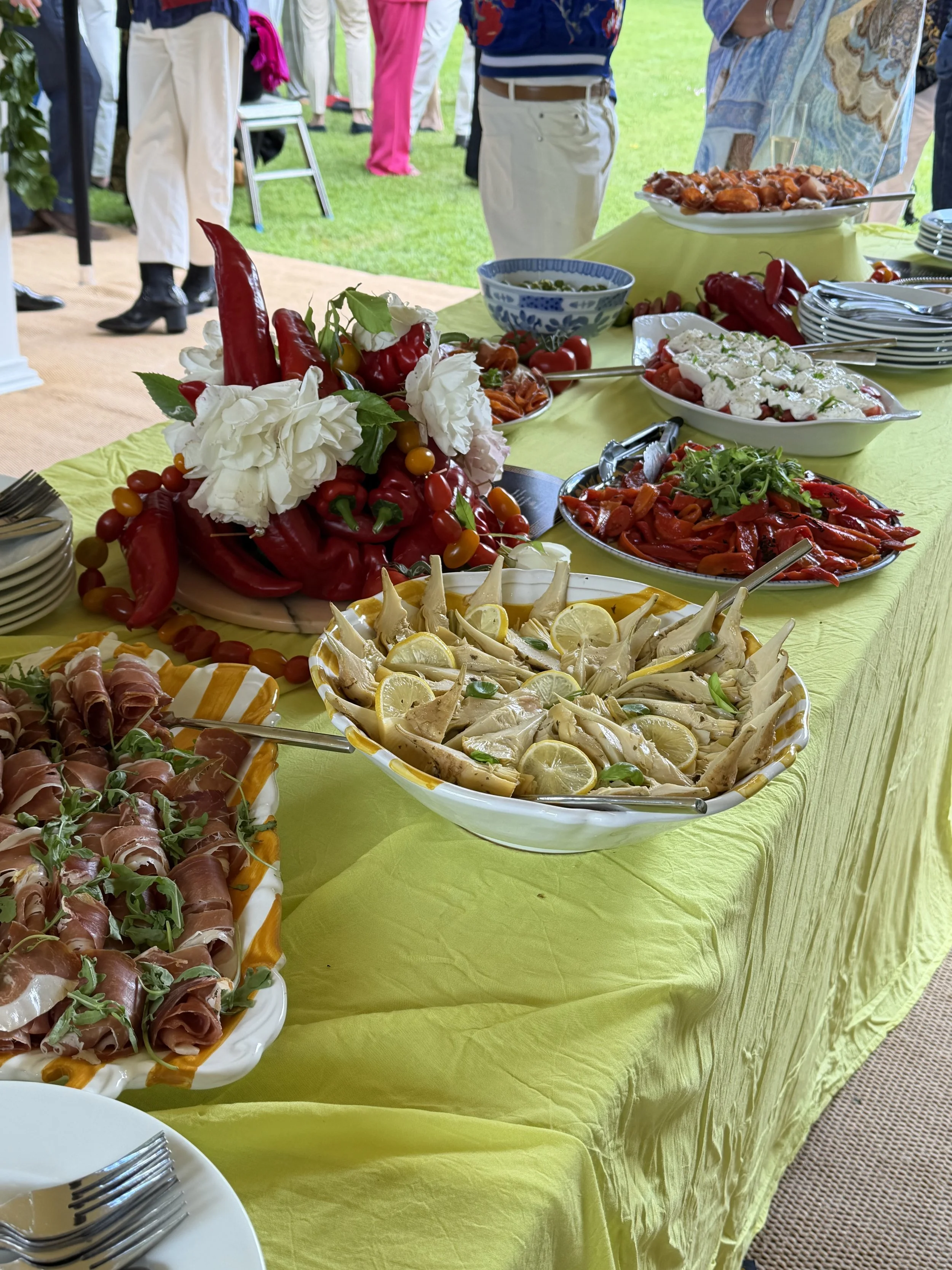 A buffet table with a yellow tablecloth features an assortment of dishes including sliced vegetables, meats, salads, and garnishes. The background shows people standing and chatting outdoors on a lawn, with some wearing colorful clothing.