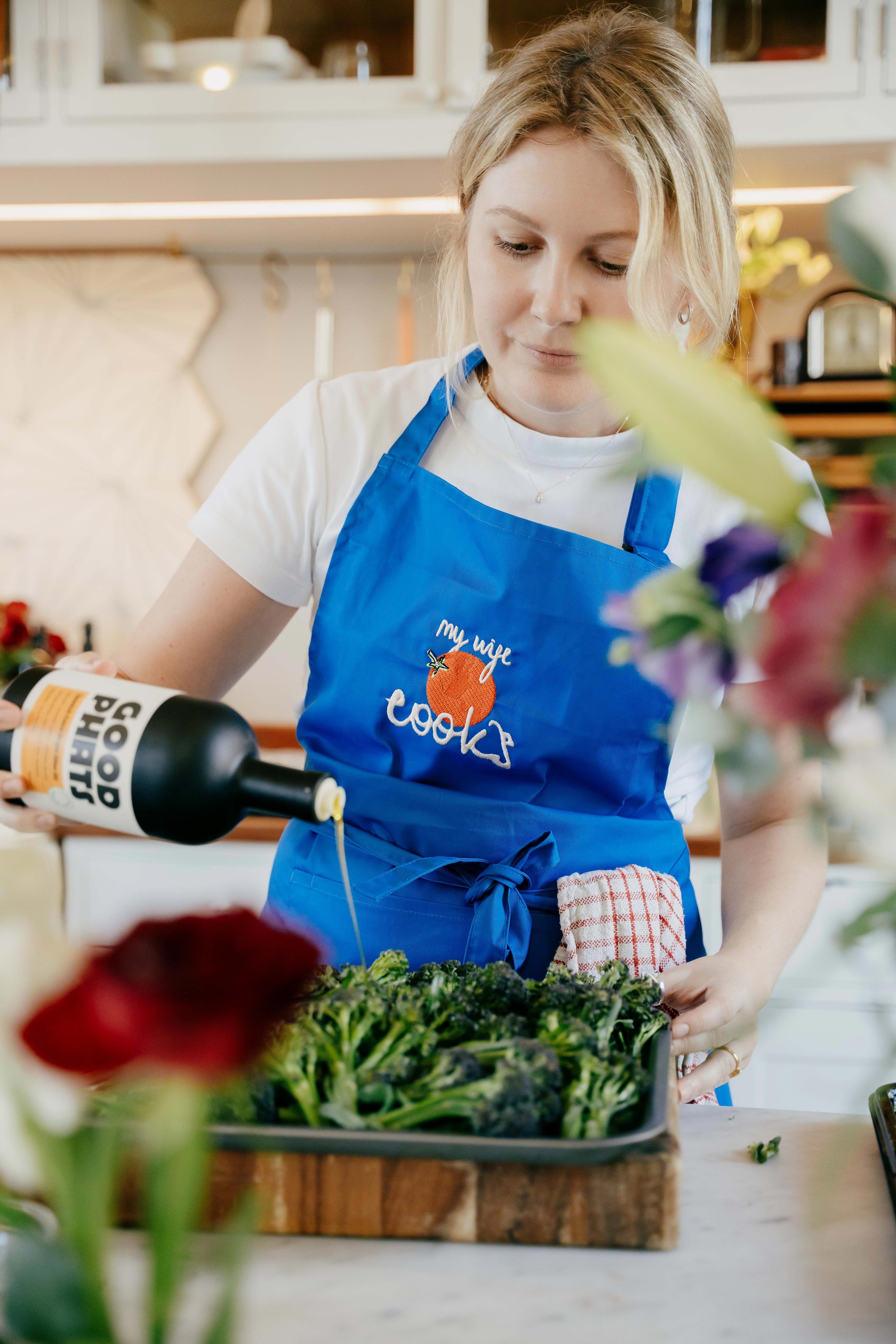 The chef in a blue apron pouring dressing from a bottle onto a salad in a kitchen.