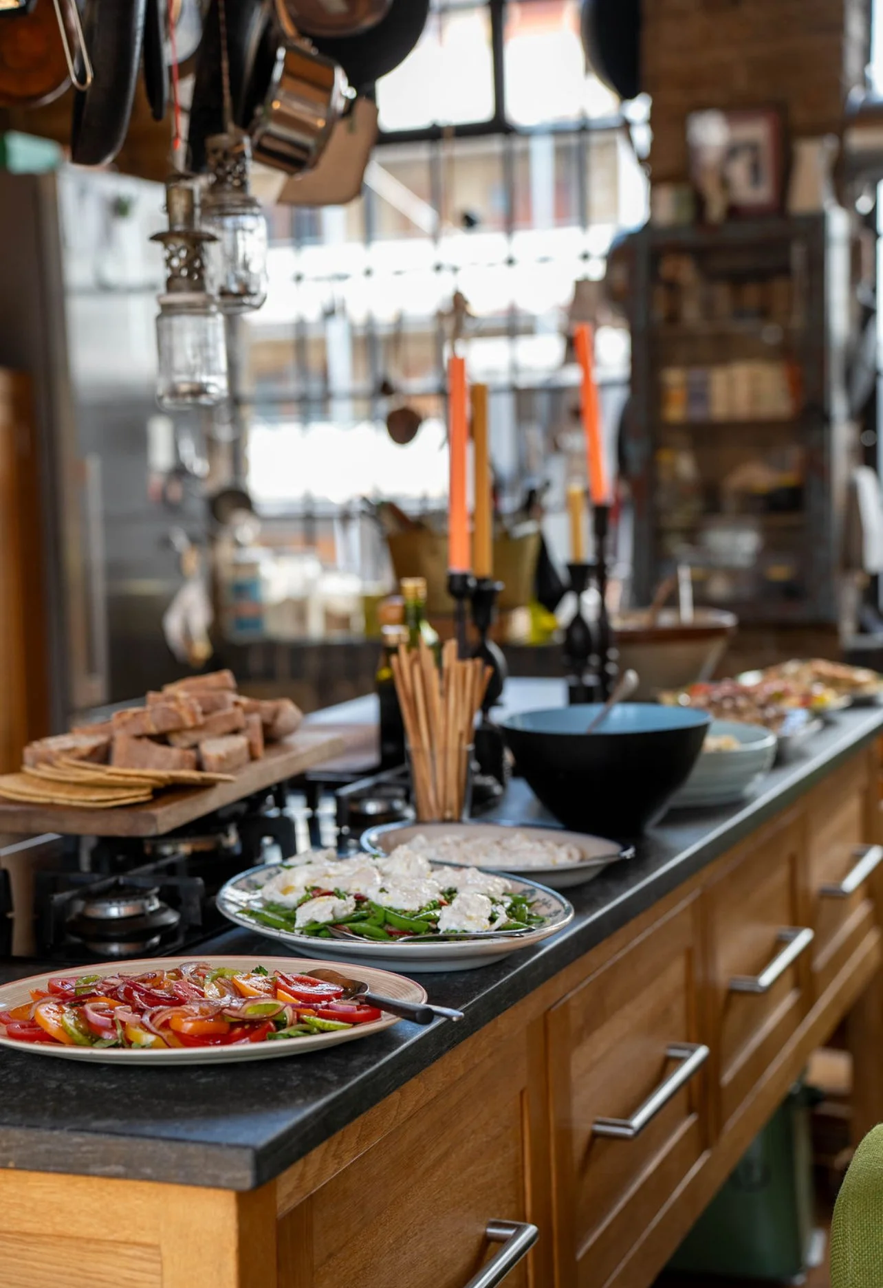 A kitchen countertop with serving dishes containing sliced tomatoes, a green salad with cheese, and a pasta dish, with a black serving bowl, utensils, and stone bread in the background.