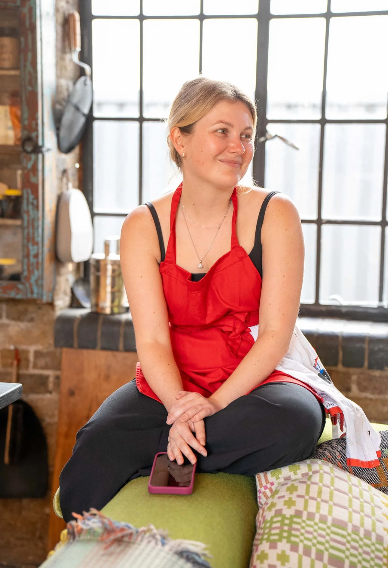 A woman with short blonde hair sits cross-legged on a sofa in a kitchen, wearing a red apron over black clothing, smiling softly and looking to her right, holding a pink smartphone on her lap, with a brick wall and large window behind her.