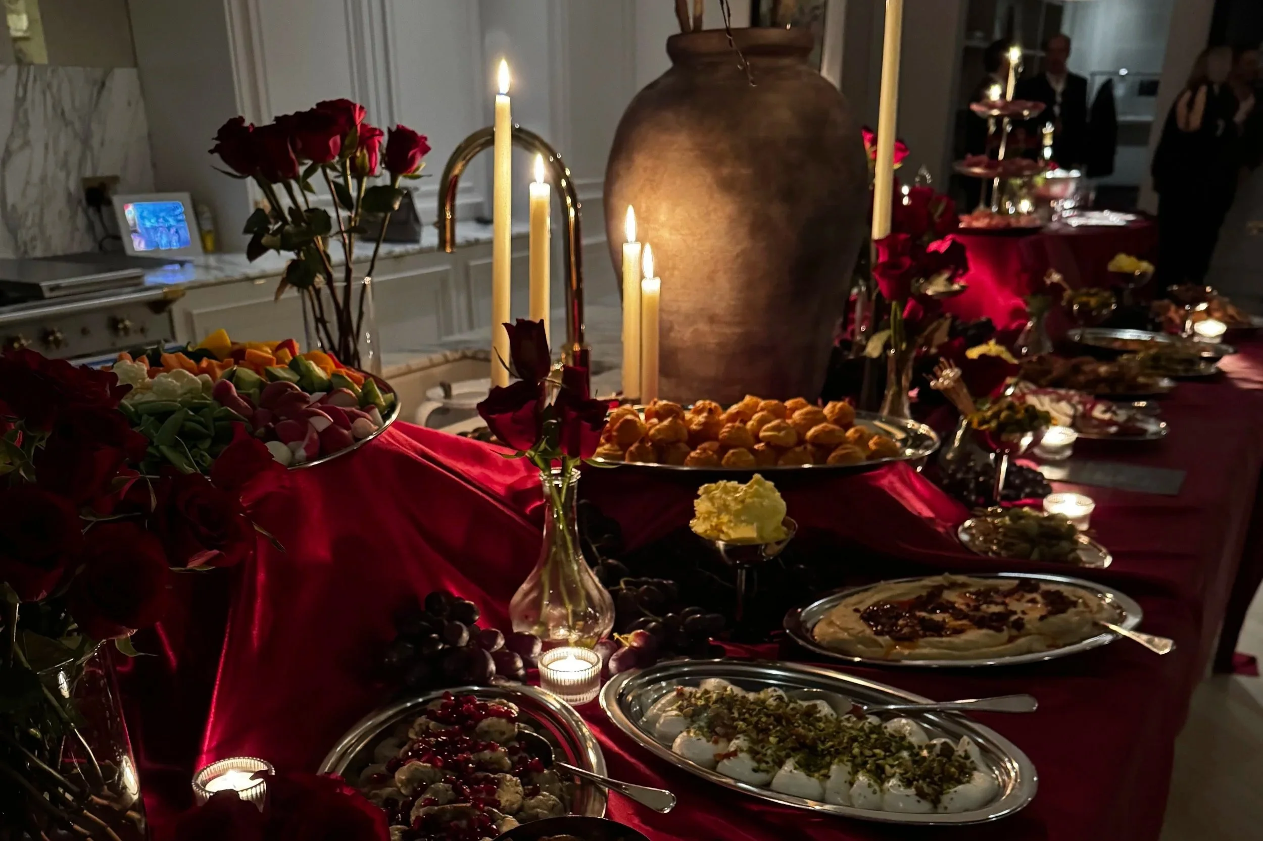 An elegant holiday buffet table decorated with red cloth, lit candles, flowers, and various desserts.
