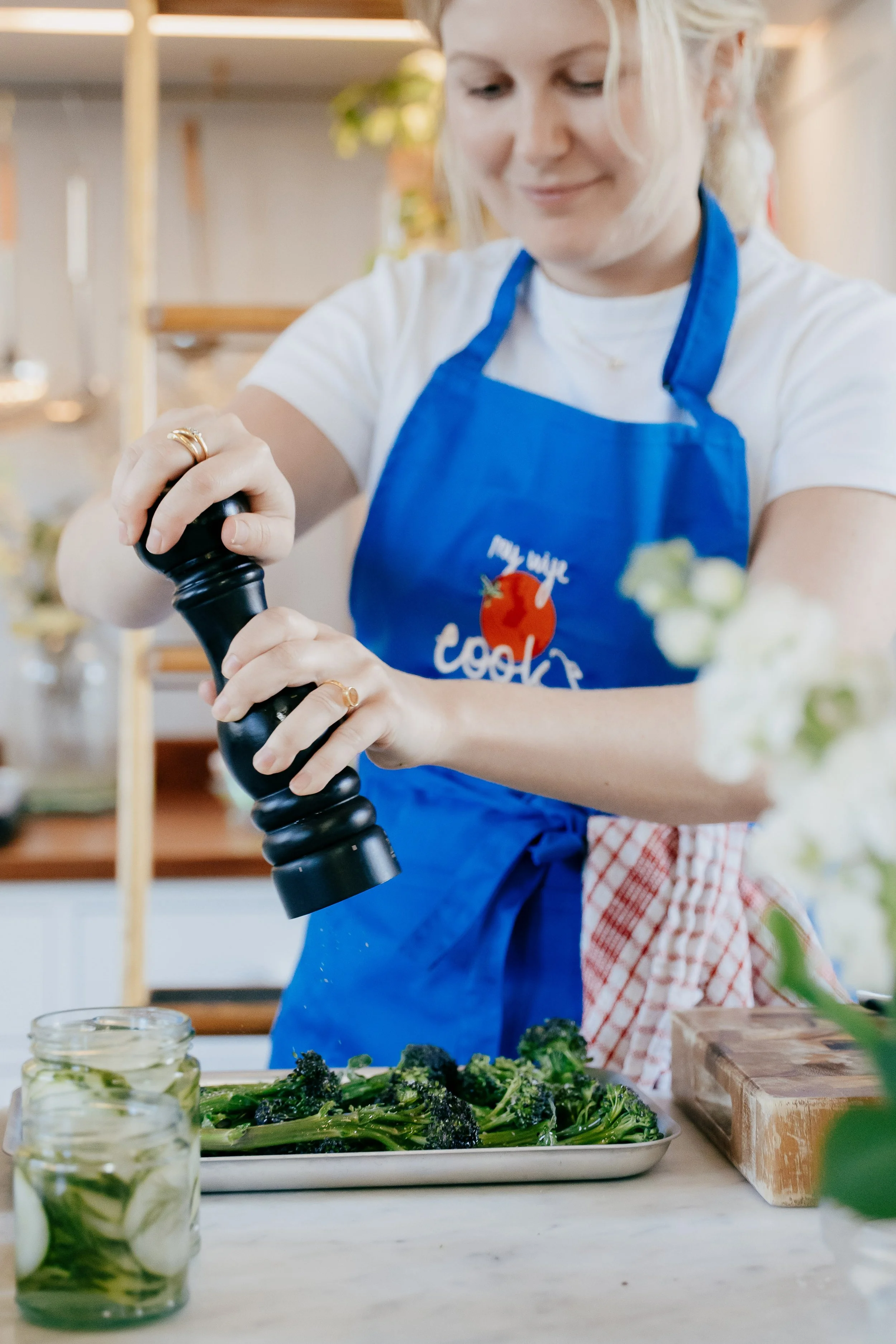 A woman wearing a white t-shirt, blue apron, and a red and white checkered towel is seasoning cooked broccolini on a kitchen counter. There are jars with preserved vegetables and a cutting board on the counter.