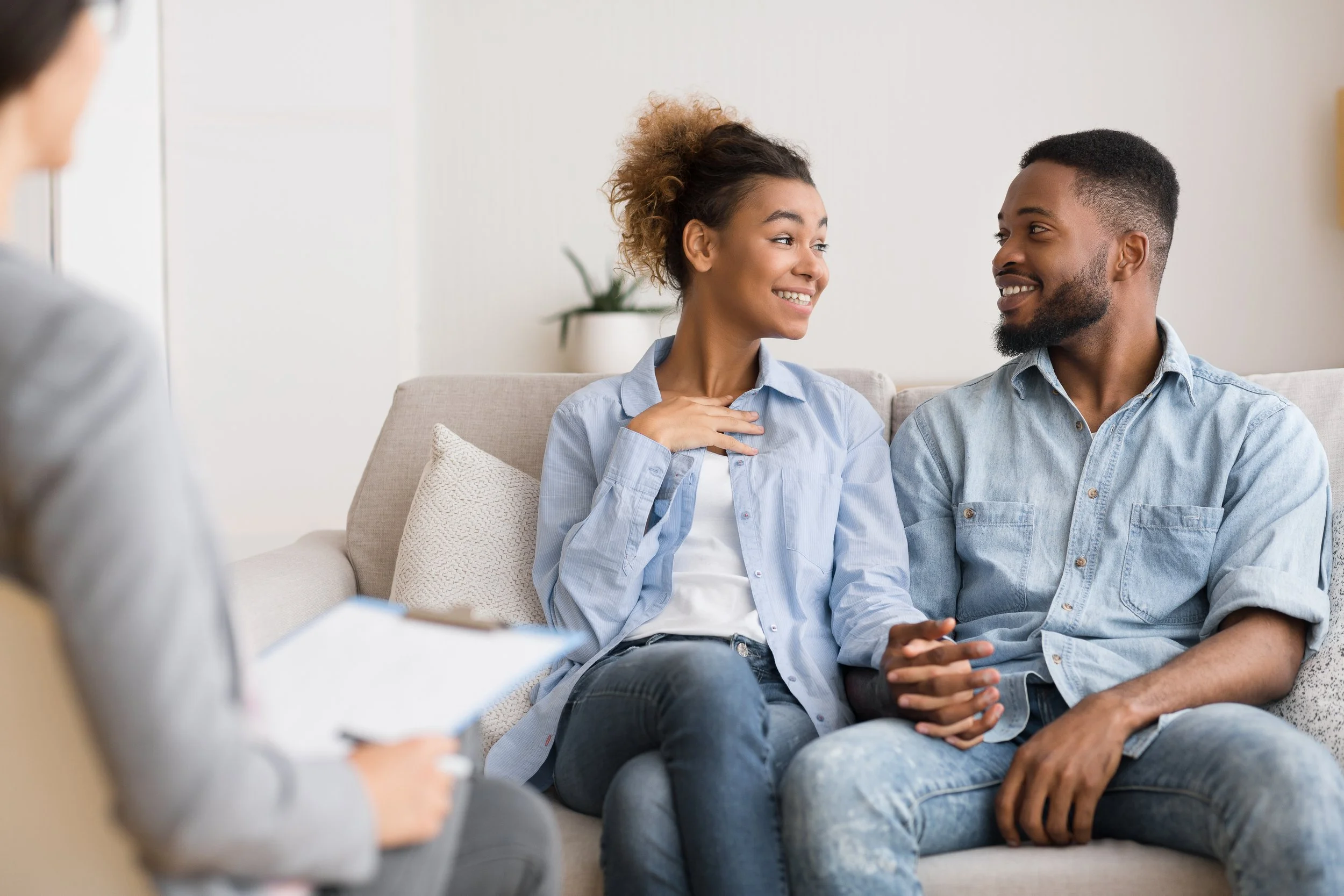 A couple sitting on a beige couch during a therapy session, smiling and holding hands while talking to a therapist holding a notebook.