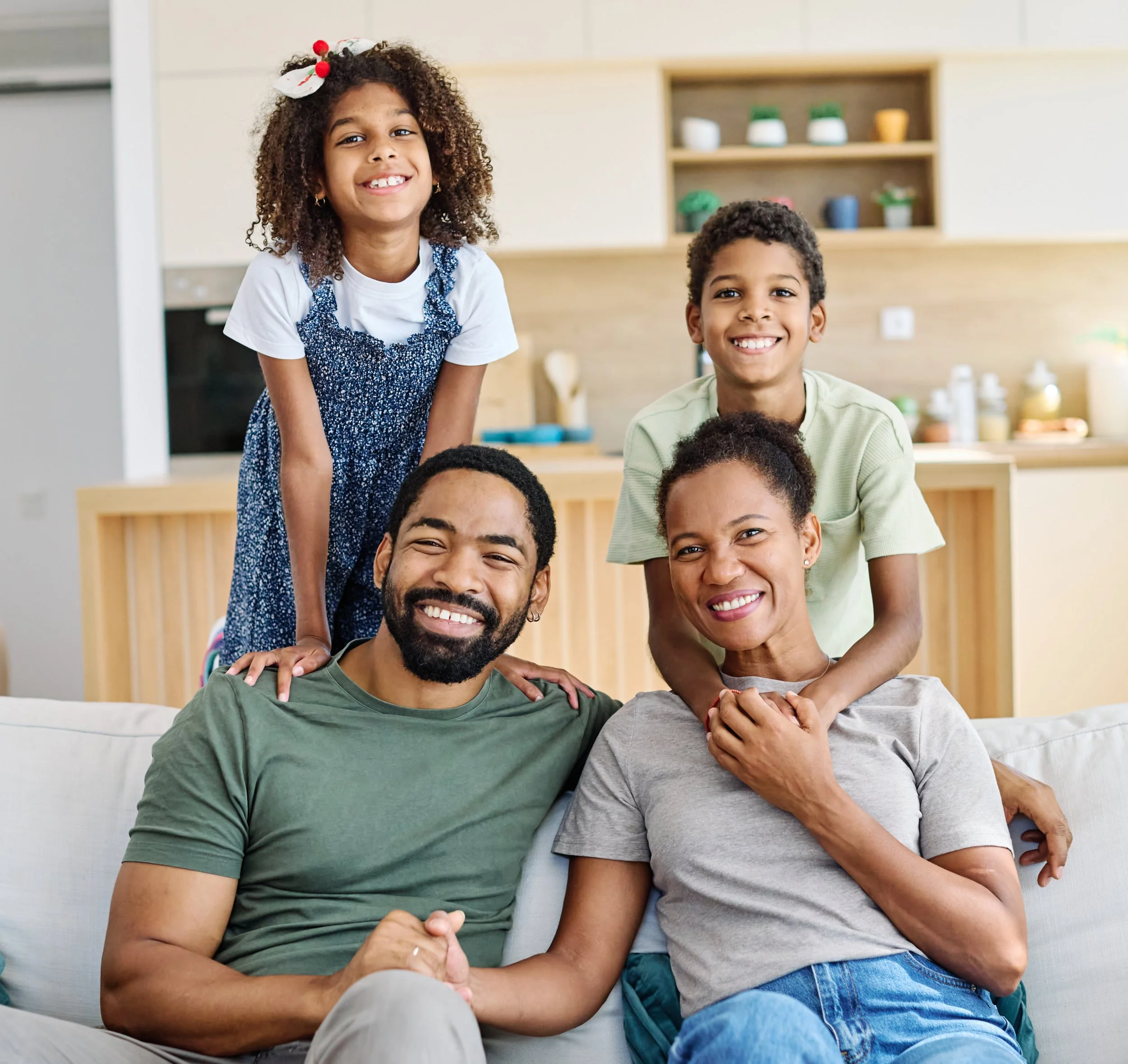A happy multigenerational African American family of four sitting on a beige couch in a modern kitchen. The adults, a man and woman, are seated close together, smiling, with the children—an older girl and a younger boy—leaning on their shoulders. The family appears joyful and close-knit.