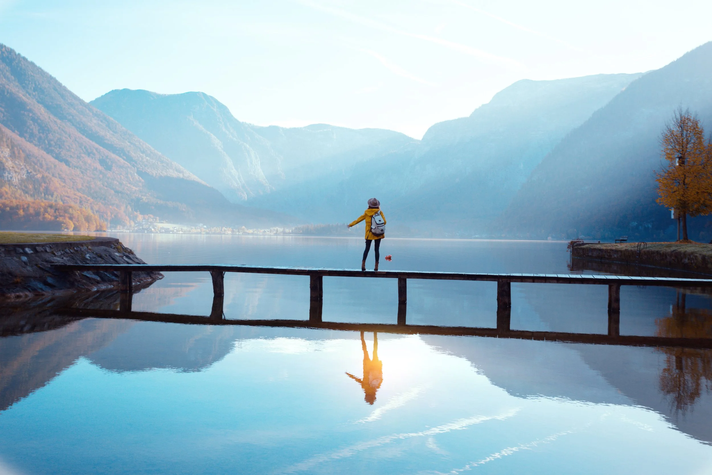 Person in yellow coat and hat standing on a wooden bridge over a calm lake, with mountains and a tree in the background, and their reflection in the water.
