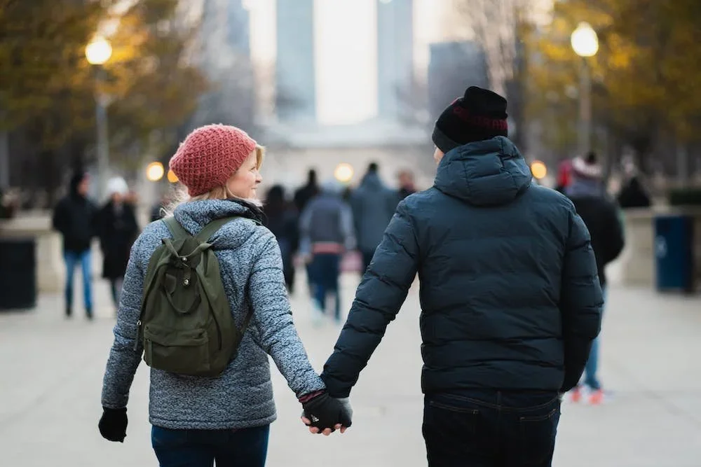 A young woman and a young man holding hands while walking in a park or city street during fall. The woman is wearing a red knit hat, a grayish-blue jacket, and a green backpack. The man is wearing a black beanie, a dark puffer jacket, and dark pants. Other people and street lamps are visible in the background.