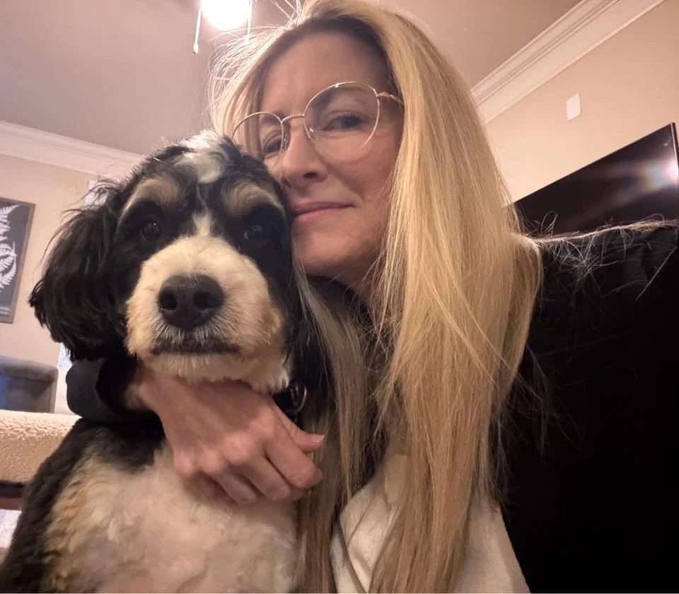 A woman with long blonde hair and glasses taking a selfie with her arm around a black and white dog.