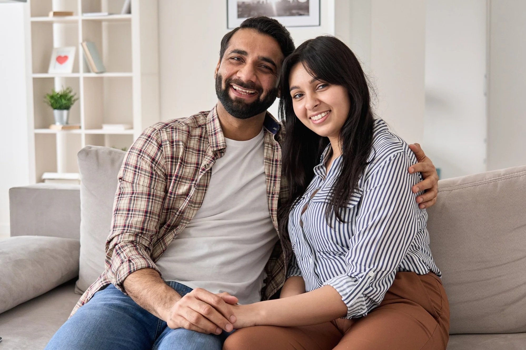 A happy couple sitting close together on a beige couch in a bright living room, smiling at the camera.