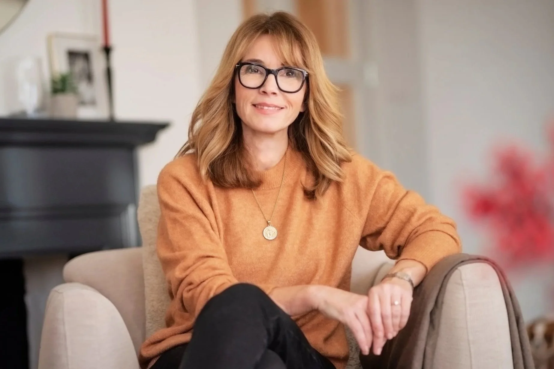 A woman with red hair, glasses, and wearing an orange sweater sitting on a beige armchair in a cozy home setting with a piano and framed pictures in the background.