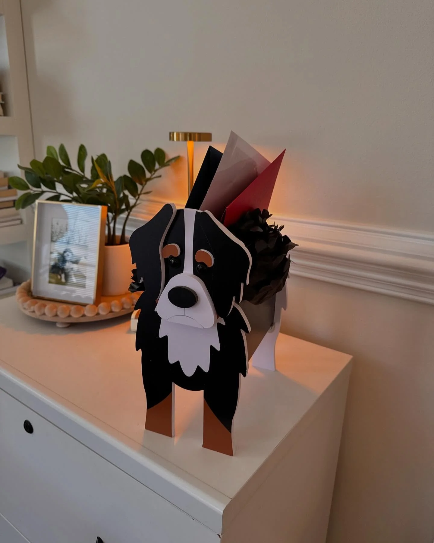 Decorative dog-shaped container with black, white, and brown paper flowers on a white cabinet, with a small plant, framed picture, and lamp in the background.