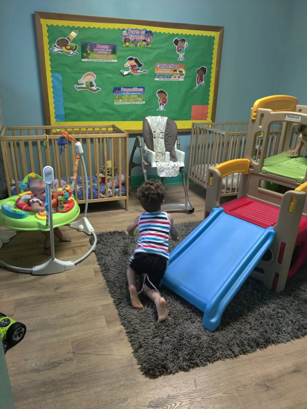 Children playing in a brightly decorated indoor play area with a small slide, a baby jumper, and cribs. A green bulletin board with educational posters about speech parts is displayed on the wall.