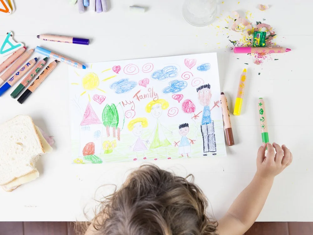 Child drawing a family picture with crayons on paper, surrounded by markers, a sandwich, and spilled crayon shavings on a white table.