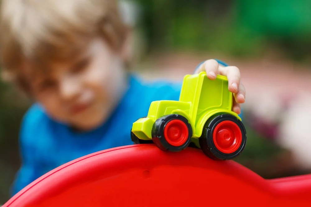 Child holding a bright green toy truck, placing it on a red surface, outdoors.