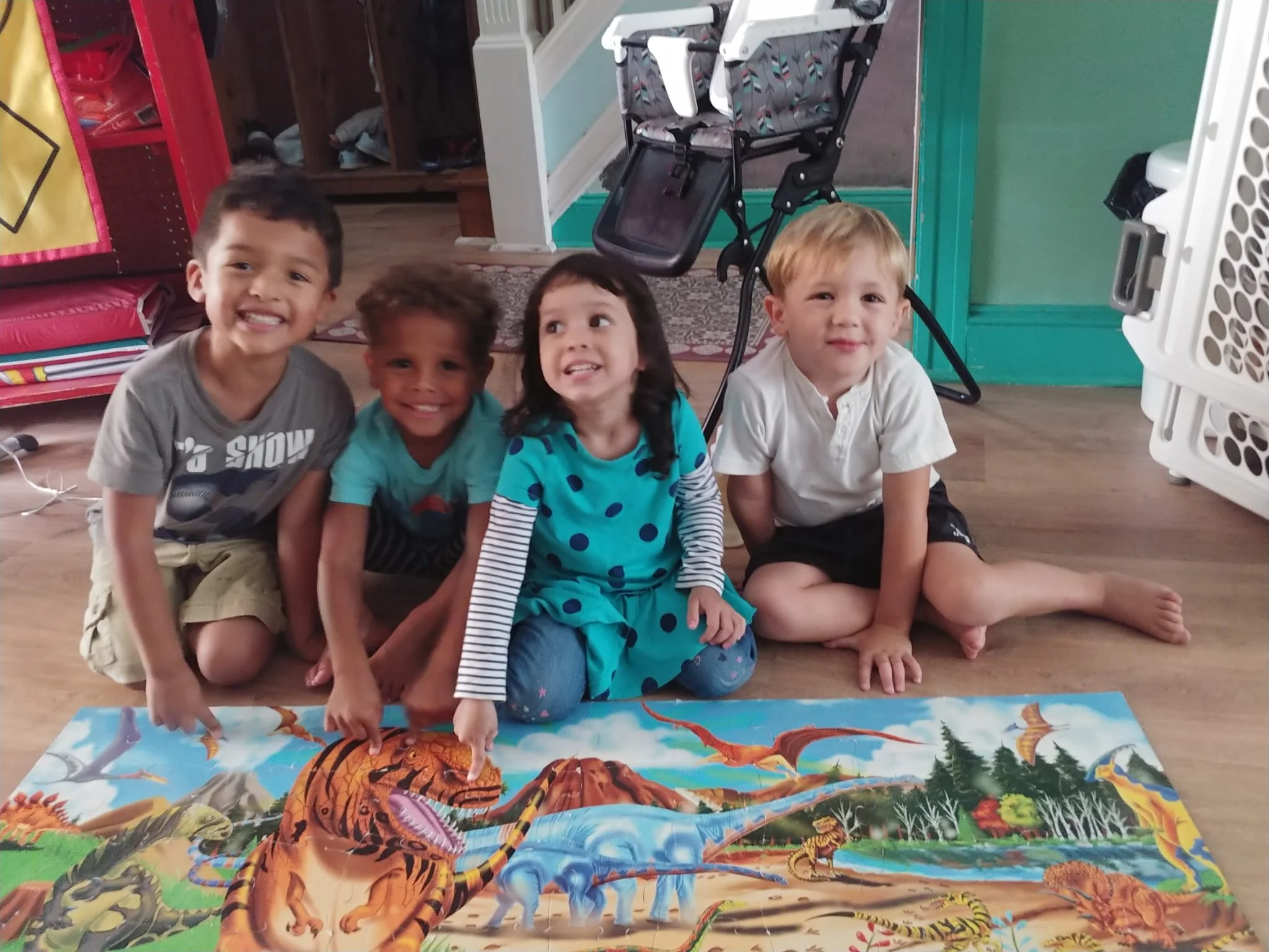Four children sitting cross-legged on a wooden floor behind a colorful dinosaur-themed puzzle. The children are smiling and looking at the camera, with two boys on the left and a girl and boy on the right.
