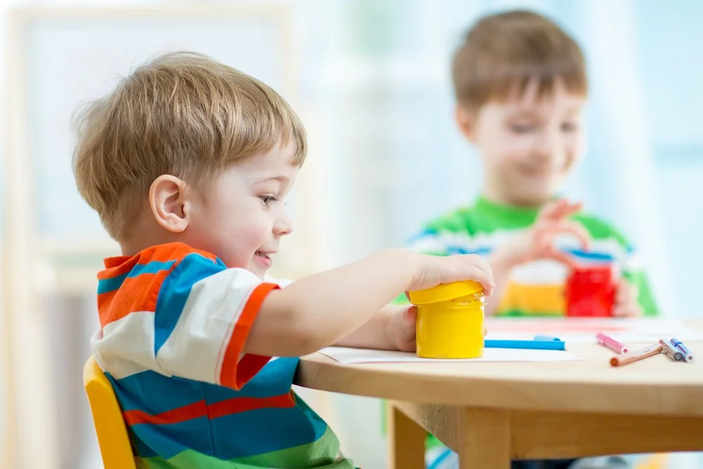 Two young boys with light brown hair playing with colorful paint jars and markers at a table.