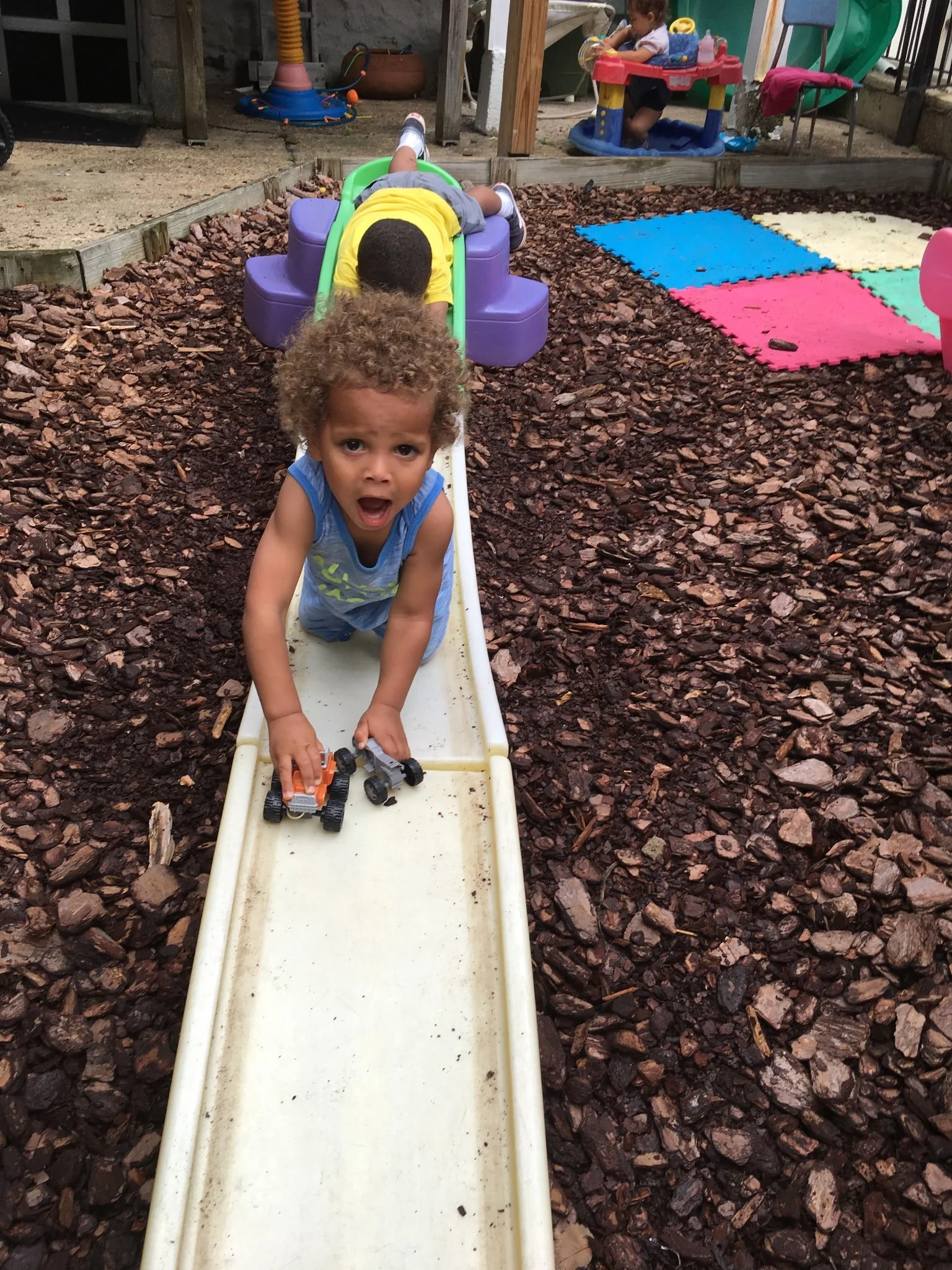 A young boy with curly hair, wearing a blue sleeveless shirt, is crawling on a small slide at a playground. He is holding toy cars in his hands and looks surprised or excited. In the background, another child is playing in a activity center, and there are colorful foam mats and various playground equipment surrounding them.