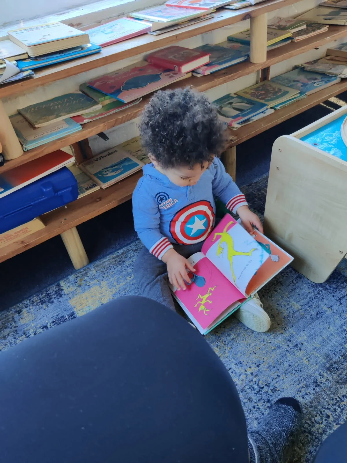 Young child sitting on the floor reading a colorful book featuring dinosaurs, with books arranged on a wooden bookshelf behind him.