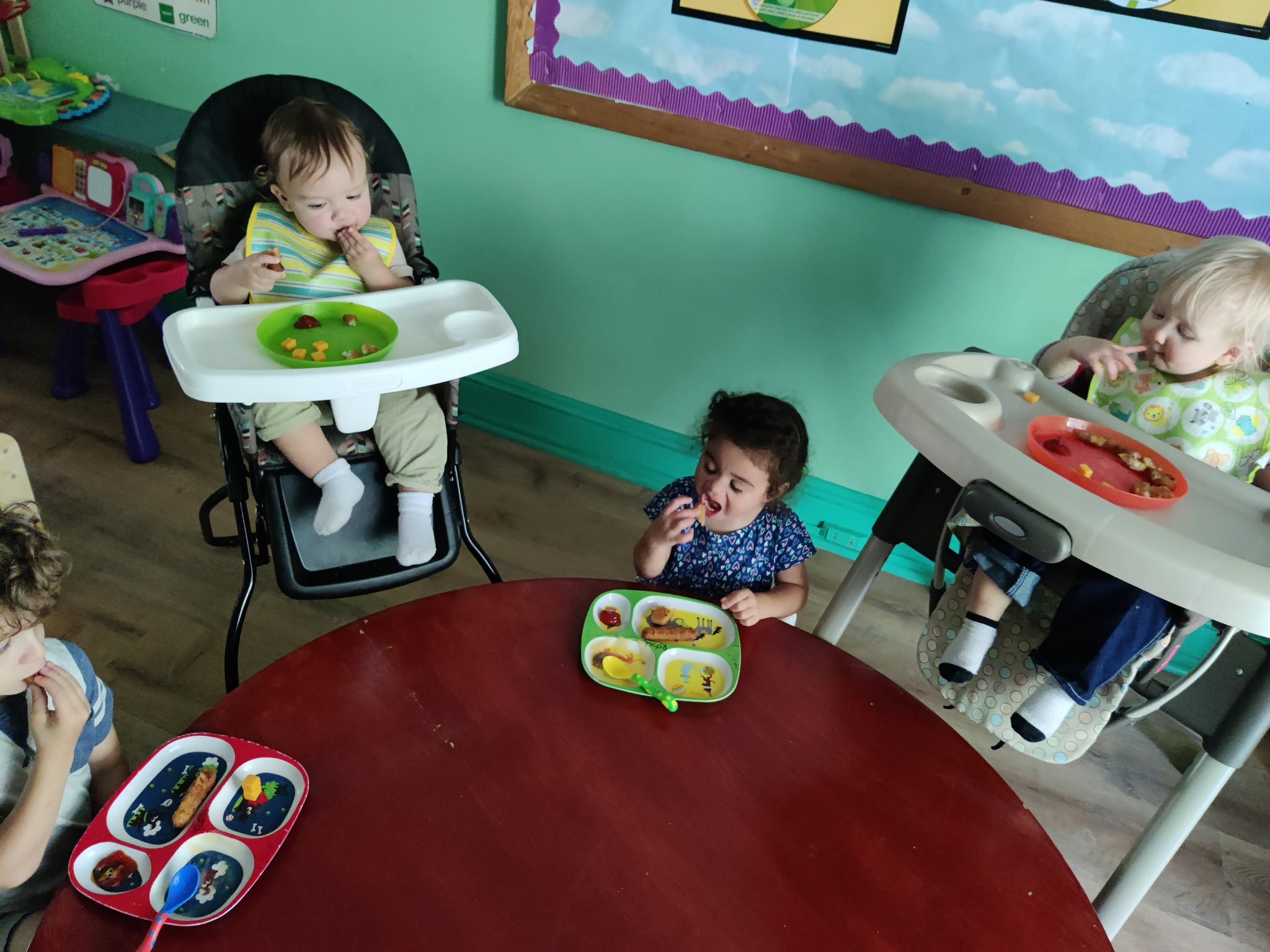 Four young children seated at a red table eating snacks. Two are in high chairs with plates of food, one girl in the middle has a divided plate with snacks, and a girl on the left is eating from a plate with cartoon characters. The room has teal walls and a bulletin board with purple trim in the background.