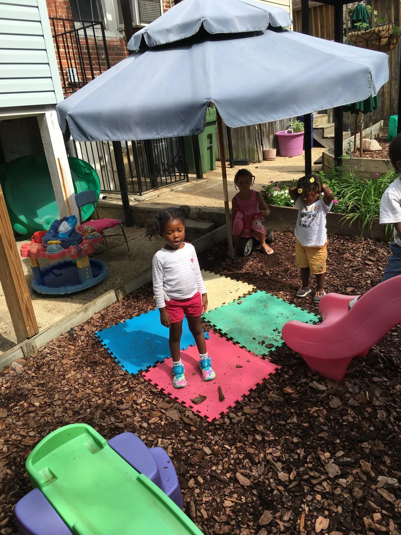 Children playing outside in a backyard under a large gray umbrella. There are colorful foam mats on the ground, a pink slide, and various toys and garden pots around.